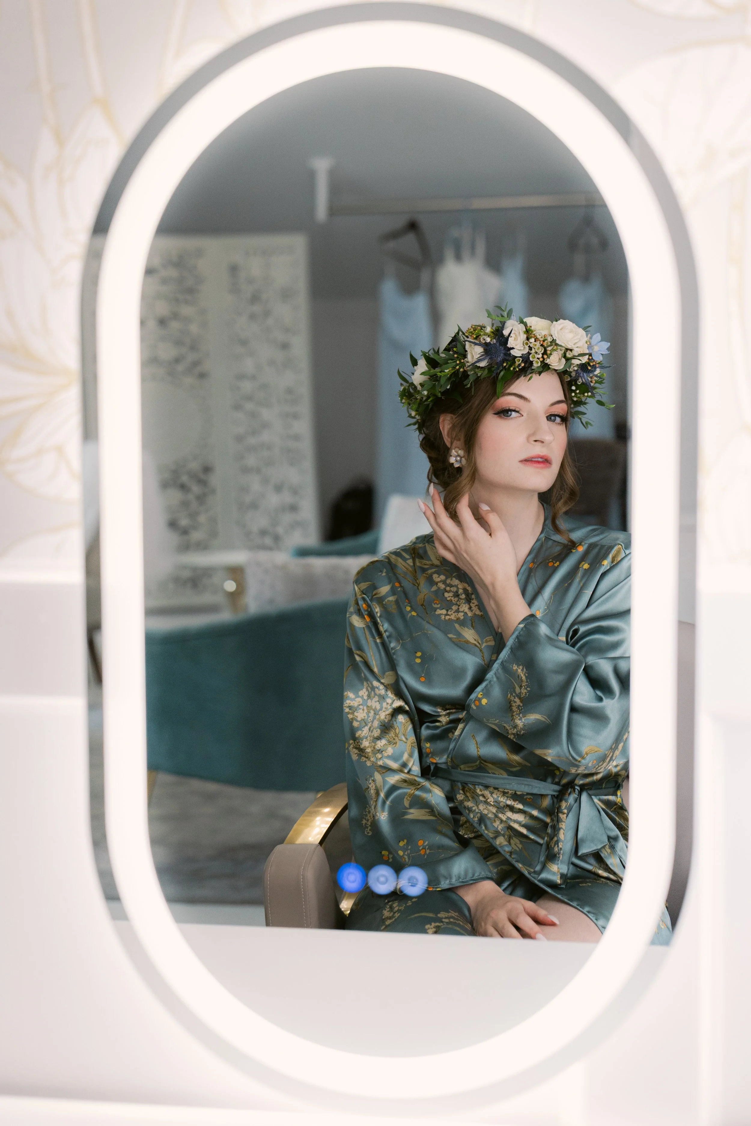 Young bride with a floral crown and silk robe sitting on a chair, looking into a mirror in the Ramble Creek bridal suite, with wedding dress hanging in the background.