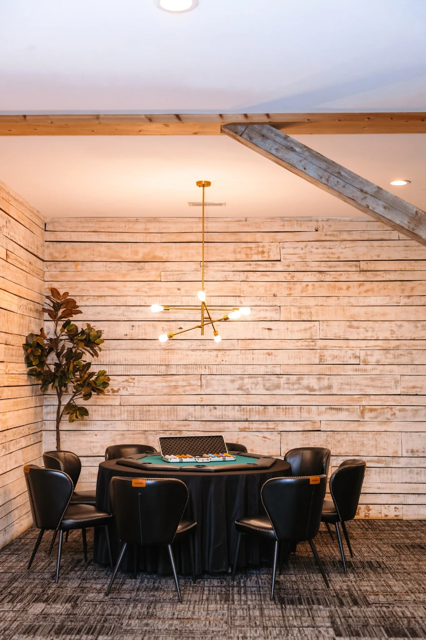 Indoor scene with a round poker table set for a game, surrounded by six black chairs, against a wooden plank wall with a leafy plant on the left, and a modern chandelier hanging from the ceiling.