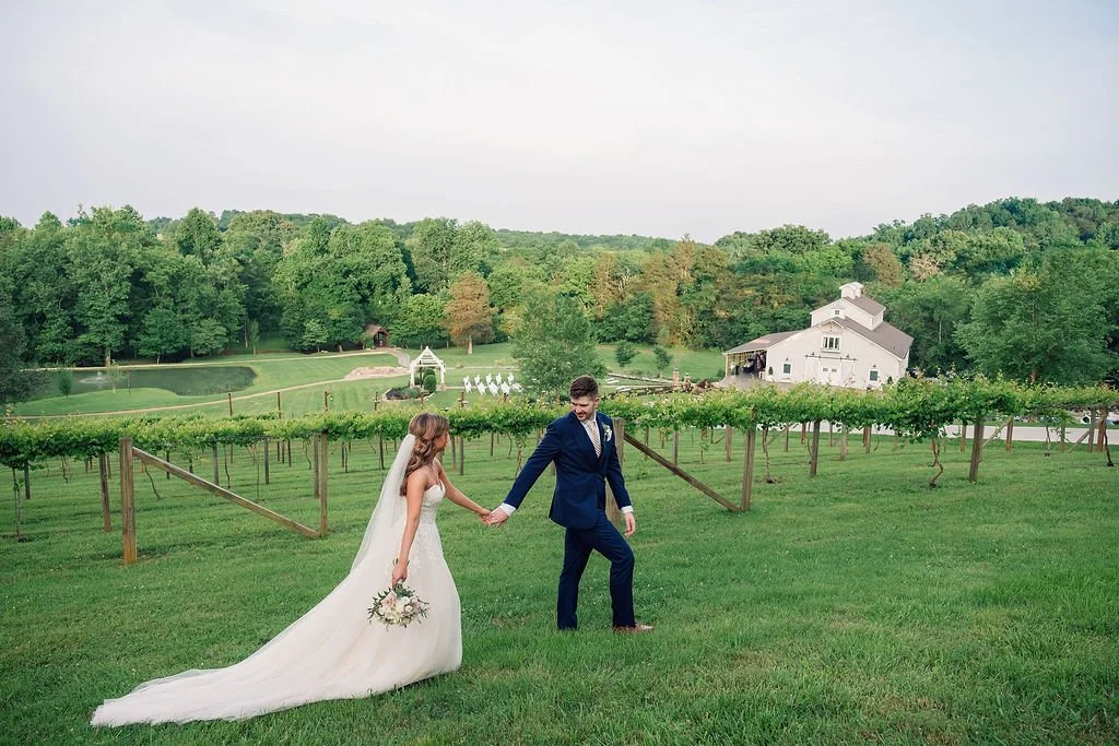 Bride and groom holding hands while walking through a grassy vineyard hillside, with a white wooden rustic venue, outdoor ceremony site, and wooded Tennessee landscape in the background