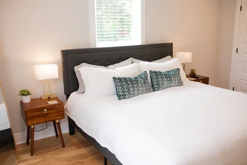 Primary bedroom inside the Villa at Ramble Creek, featuring white bedding, patterned accent pillows, wooden nightstands, bedside lamps, and a bright window.
