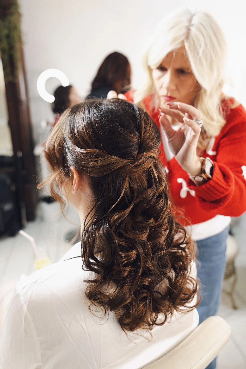 Bride’s half-up curled wedding hairstyle shown from behind as a stylist adds final touches, highlighting soft volume and romantic modern waves