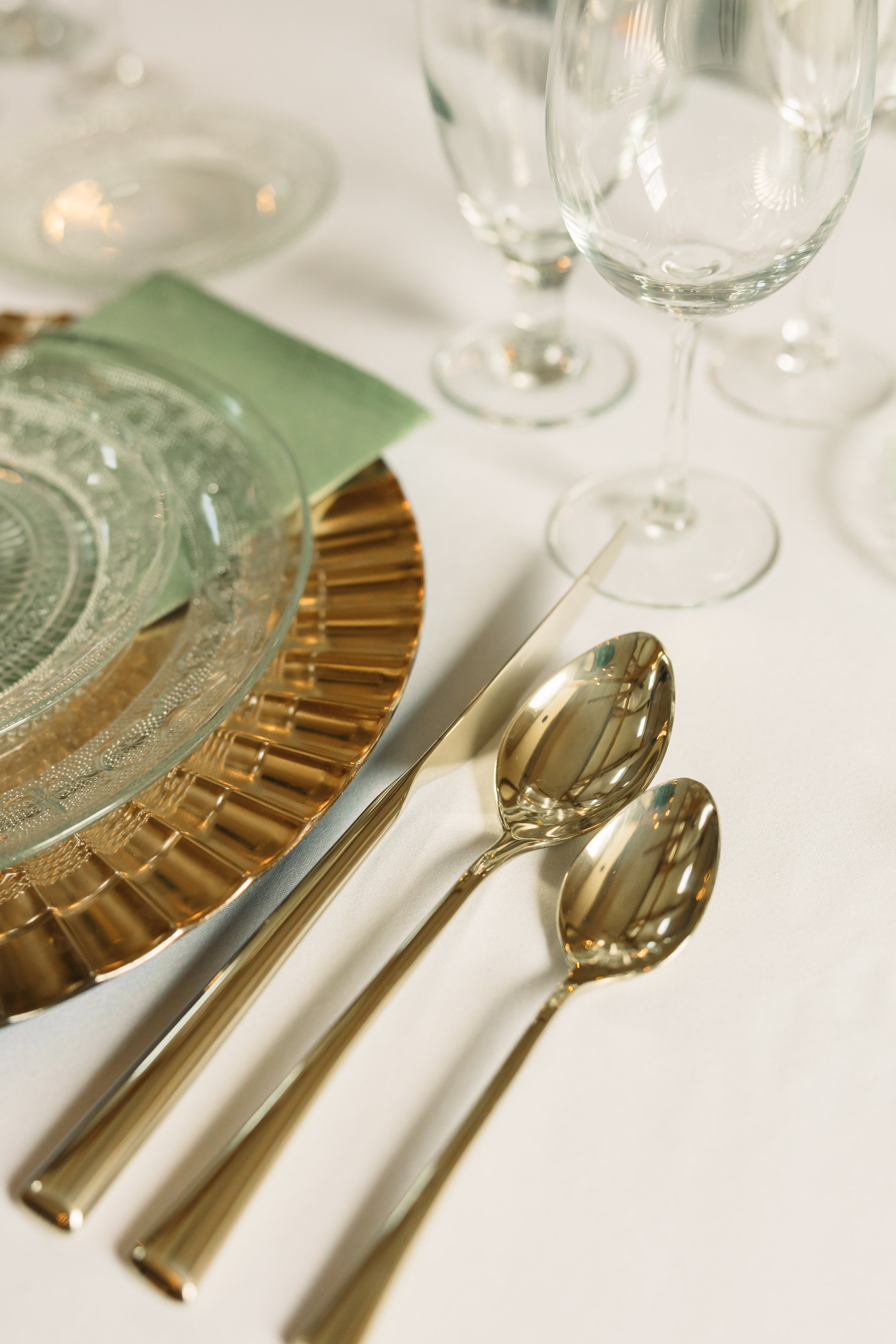 Elegant wedding reception place setting at Ramble Creek with gold flatware, glass plates, sage napkins, and polished glassware.