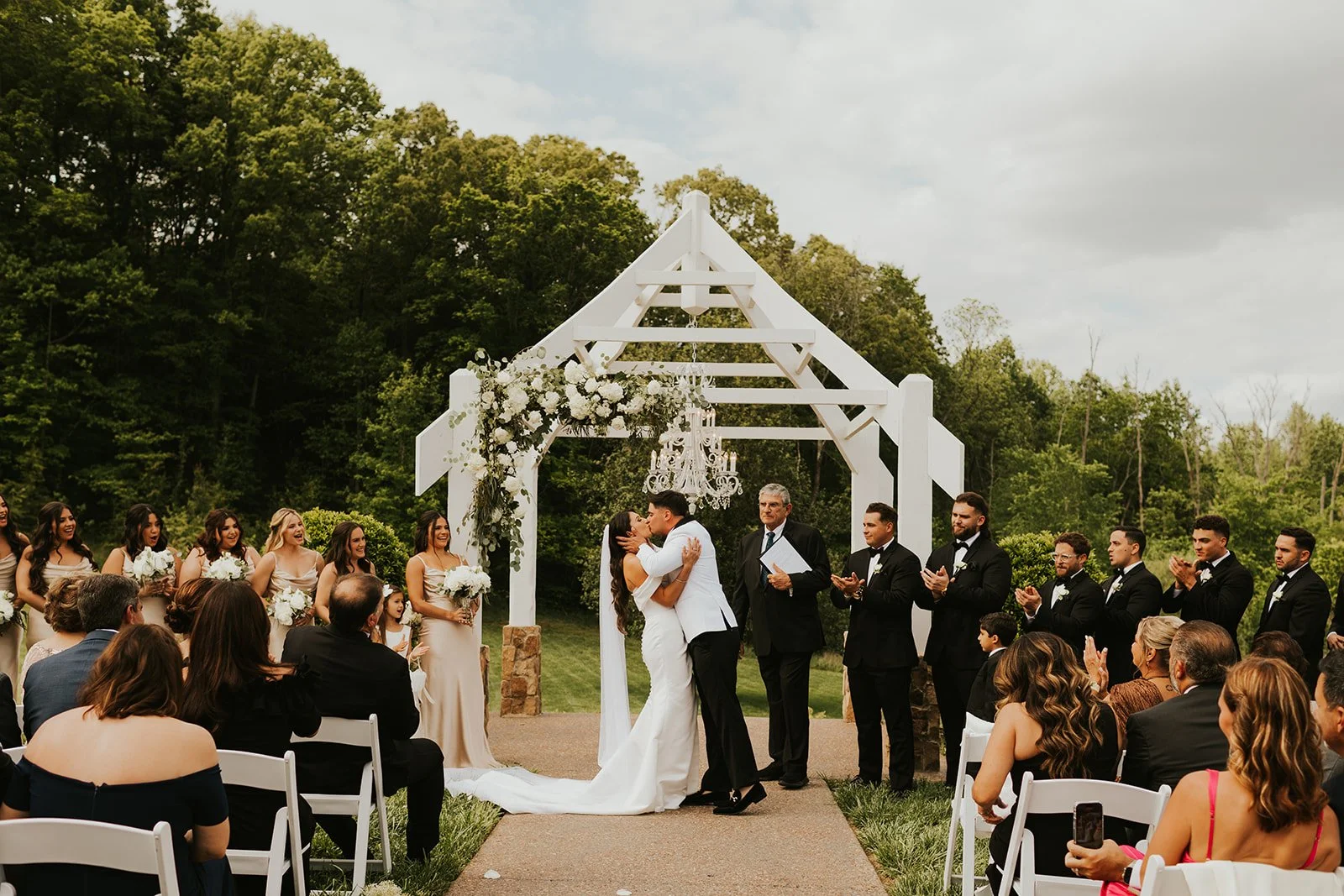 A fall/winter TN wedding ceremony outdoors with a bride and groom kissing under a white wooden arch decorated with flowers, surrounded by guests and bridesmaids and groomsmen.