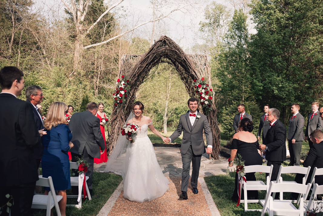 A bride and groom holding hands, walking down the aisle at an outdoor wedding ceremony, with guests on both sides and a decorative archway behind them.