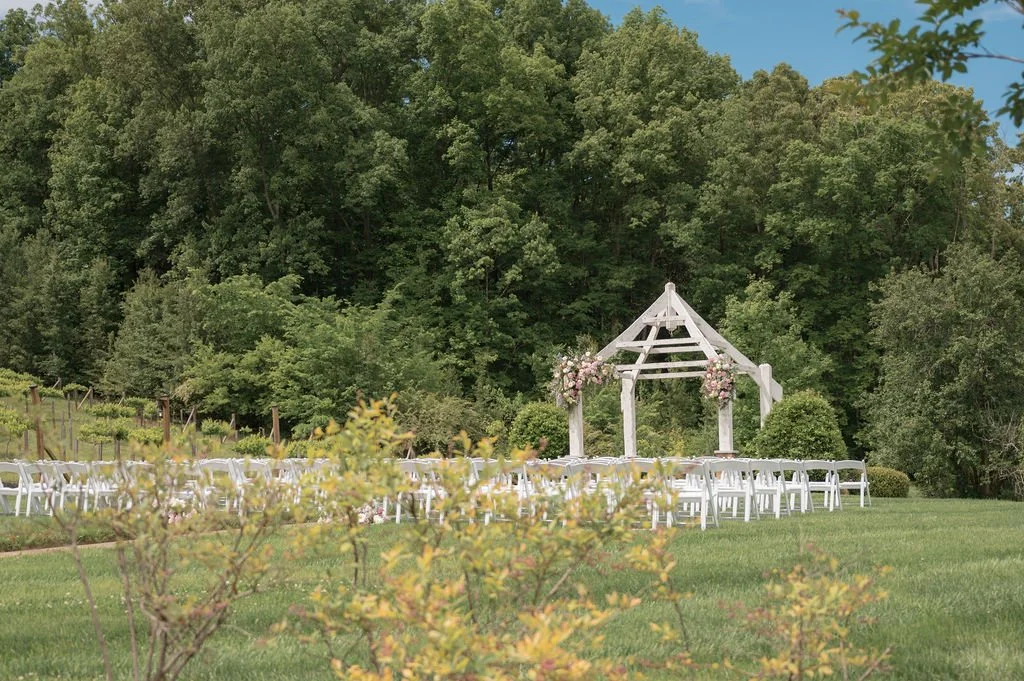 Tennessee outdoor wedding ceremony setup with a white wooden arch decorated with pink flowers and white folding chairs arranged on a grassy lawn in front of a wooded area with green trees.