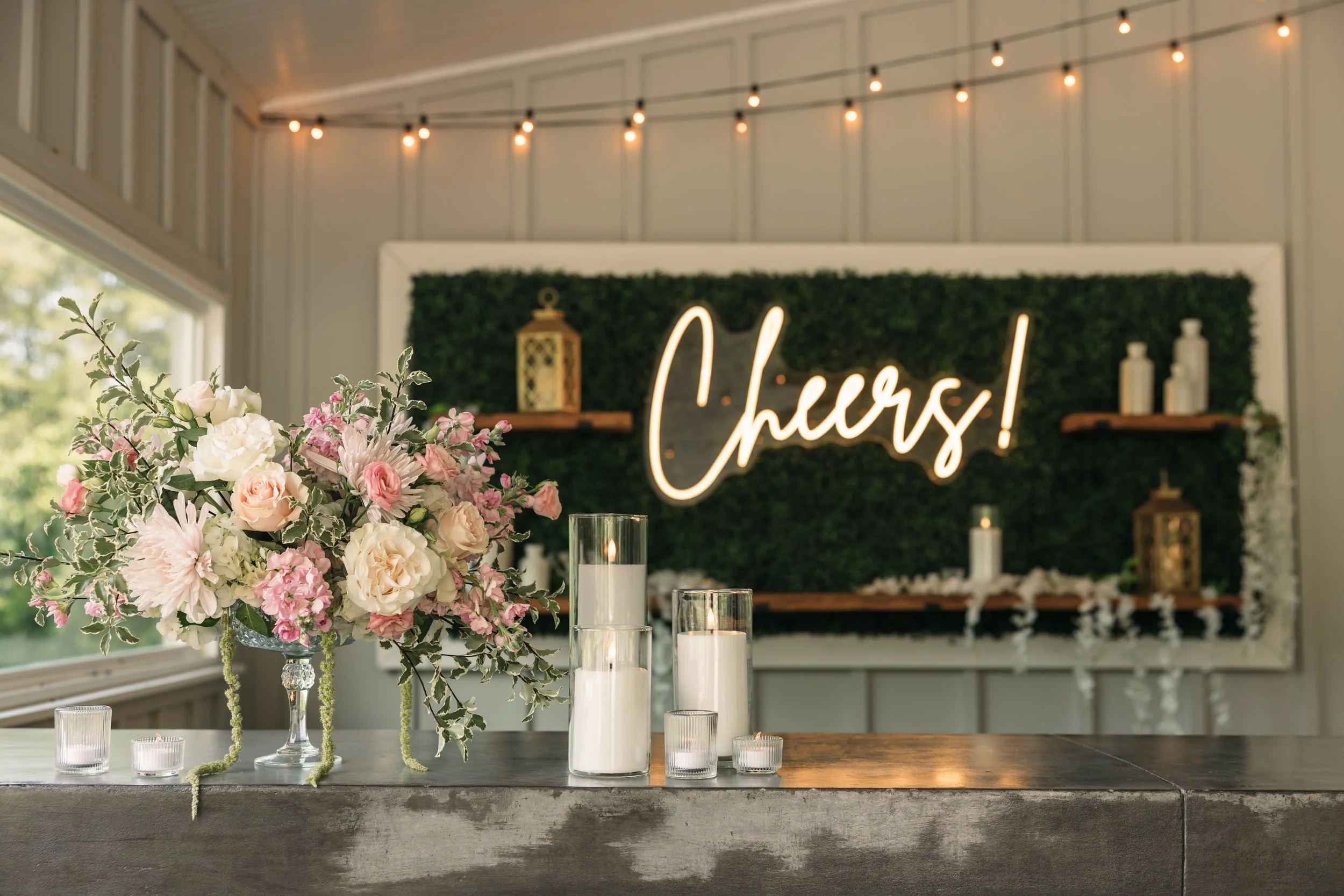Pastel floral arrangement and candlelit bar display for cocktail hour at Ramble Creek wedding venue in Tennessee with a glowing Cheers sign.