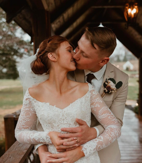 A bride and groom sharing a kiss on their wedding day outside a rustic covered bridge with trees in the background at Ramble Creek East Tennessee wedding venue.
