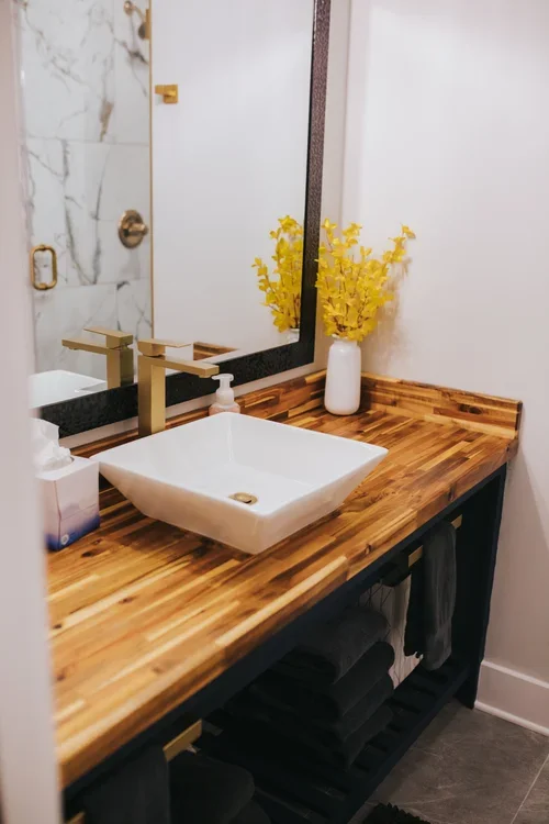 Bathroom vanity with a wooden countertop, white vessel sink, gold faucet, white soap dispenser, tall white vase with yellow flowers, large mirror, towel rack with black towels, and a reflection of the bathroom door and shower in the mirror.