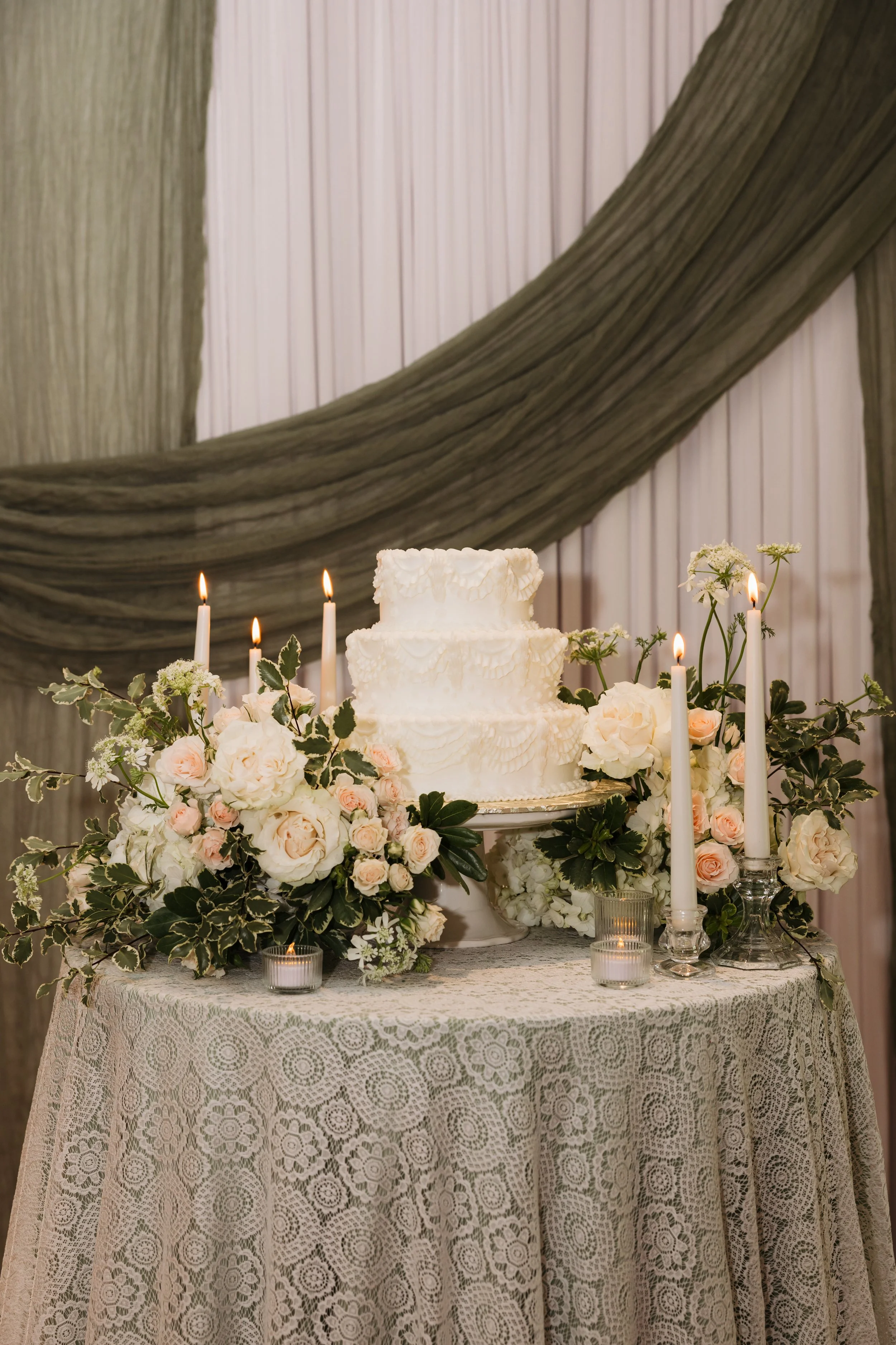 A tiered white wedding cake surrounded by white and blush roses, greenery, lit candles, and small votive candles on a lace-covered table with a dark, draped fabric background.