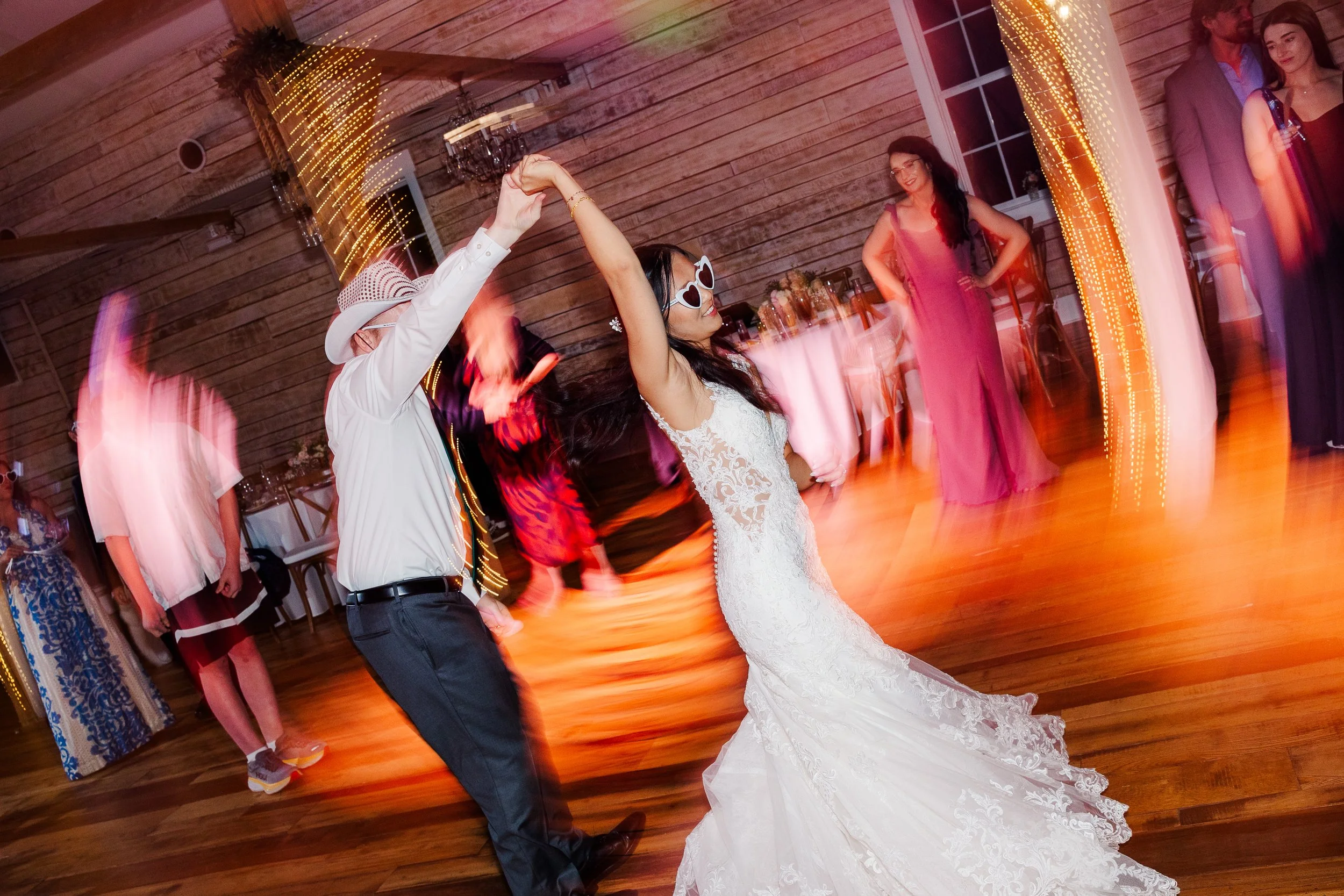 A bride and groom dancing at indoor wedding reception with guests watching in the background, some wearing sunglasses and colorful dresses, in a lively wooden wedding venue with string lights.