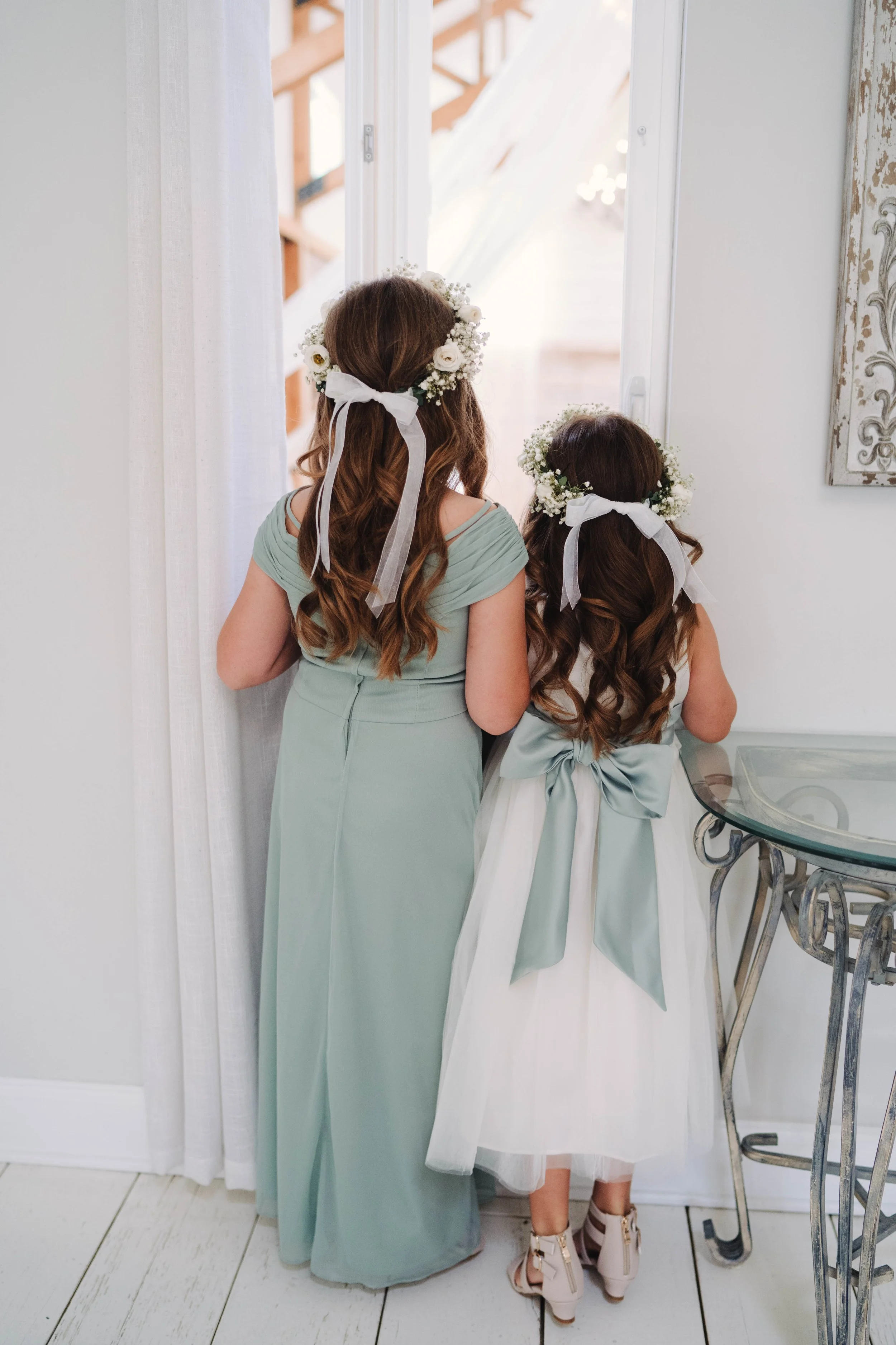 Two girls, one older and one younger, with long brown hair and floral crowns, facing a window. The older girl wears a light green dress, and the younger girl wears a white dress with a large sash. They are indoors, near a white curtain, with a metal 