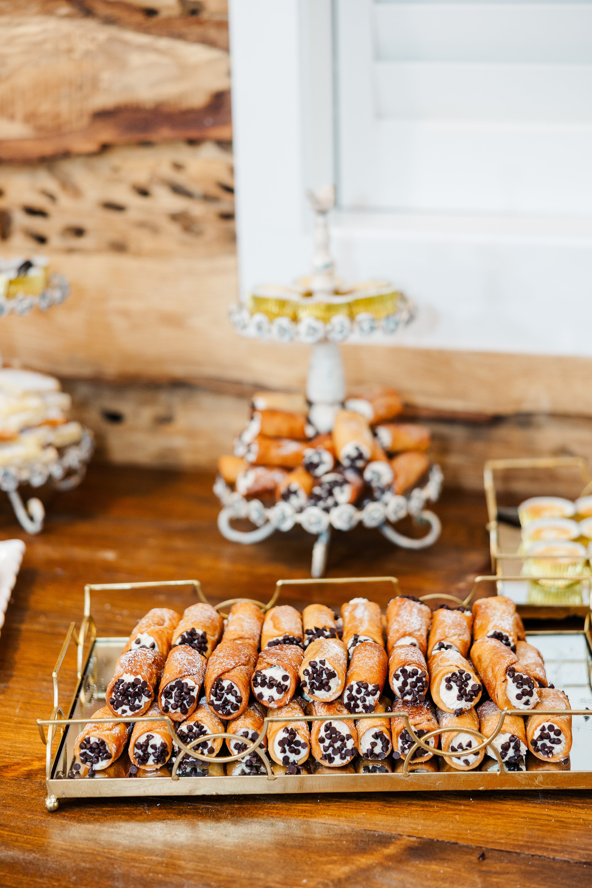 Tray of cannoli with chocolate chips on a wooden table, with a decorative stand in the background. Served as wedding desserts.