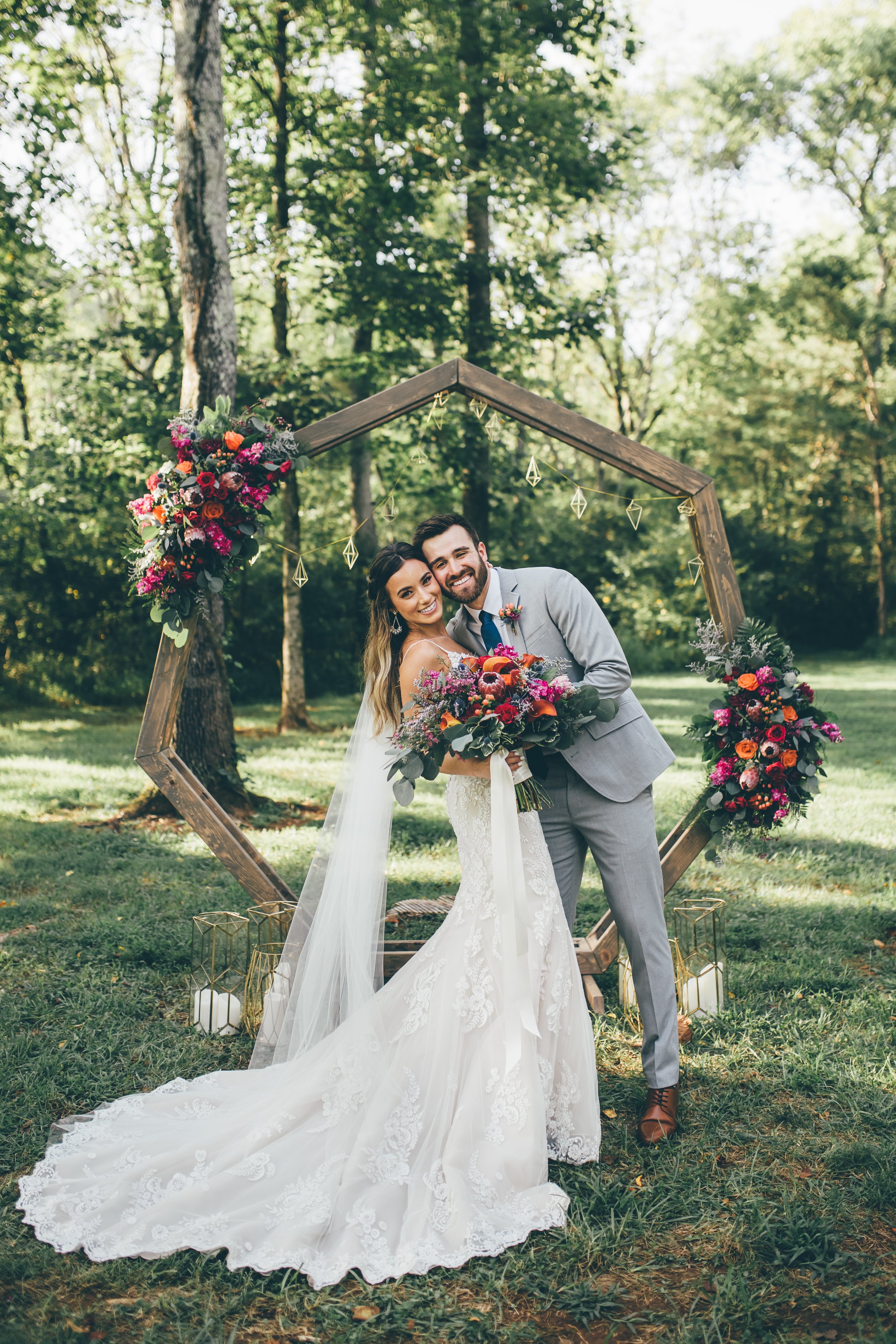 A bride and groom smile and pose together at an outdoor wedding ceremony at Ramble Creek all-inclusive wedding venue, standing in front of a geometric wooden arch decorated with colorful flowers in a wooded area.