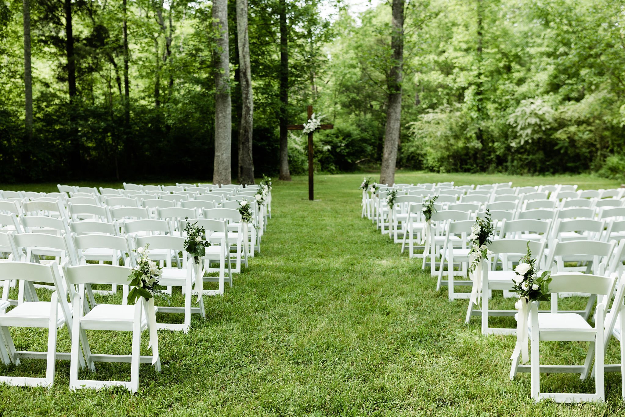 Outdoor wedding ceremony setup with white chairs decorated with flowers, a wooden cross, and a wooded background at Ramble Creek destination wedding venue in Tennessee.