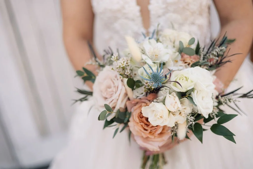 Bride holding a soft peach and ivory wedding bouquet with roses, ranunculus, blue thistle, eucalyptus greenery, and delicate filler flowers.