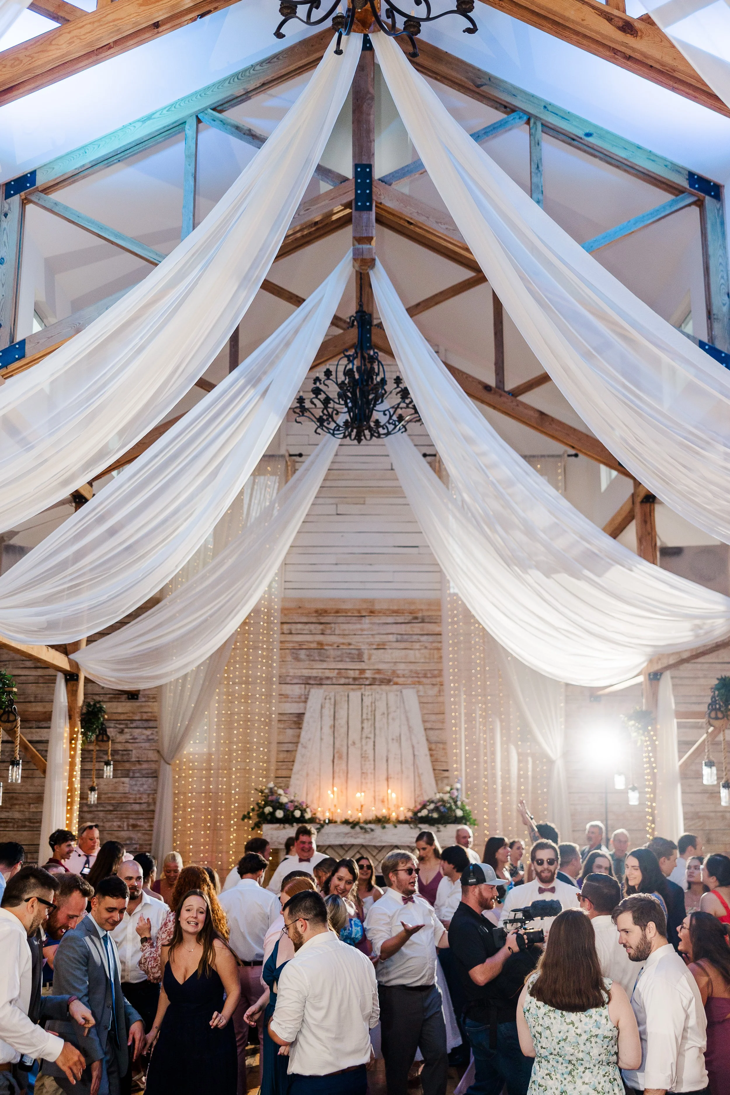 Wedding guests dancing beneath white ceiling drapes, wood beams, chandeliers, and warm string lights inside a reception venue with vaulted ceiling