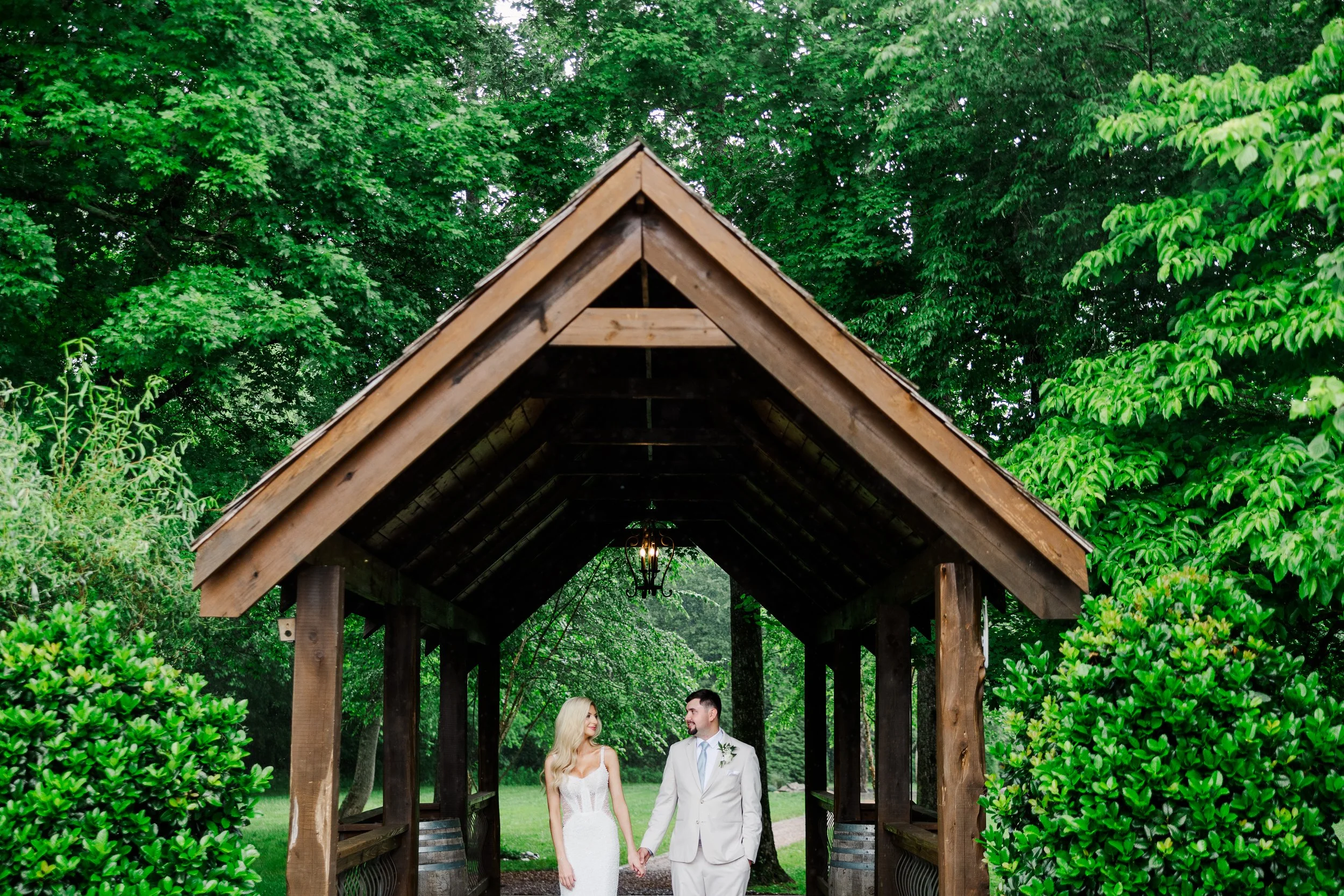 Bride and groom in lush green forest, holding hands under brown covered bridge