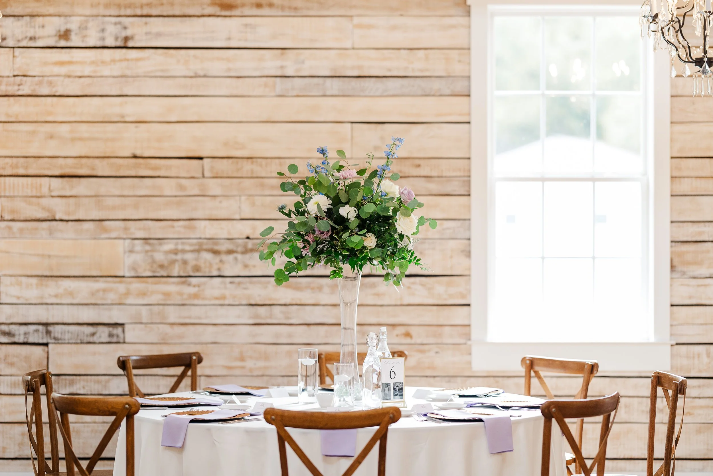 A round table with a white tablecloth set for a formal event, decorated with a tall flower centerpiece with white, pink, and blue flowers and greenery. Surrounding the table are wooden chairs with curved backs. In the background, a wall made of horiz