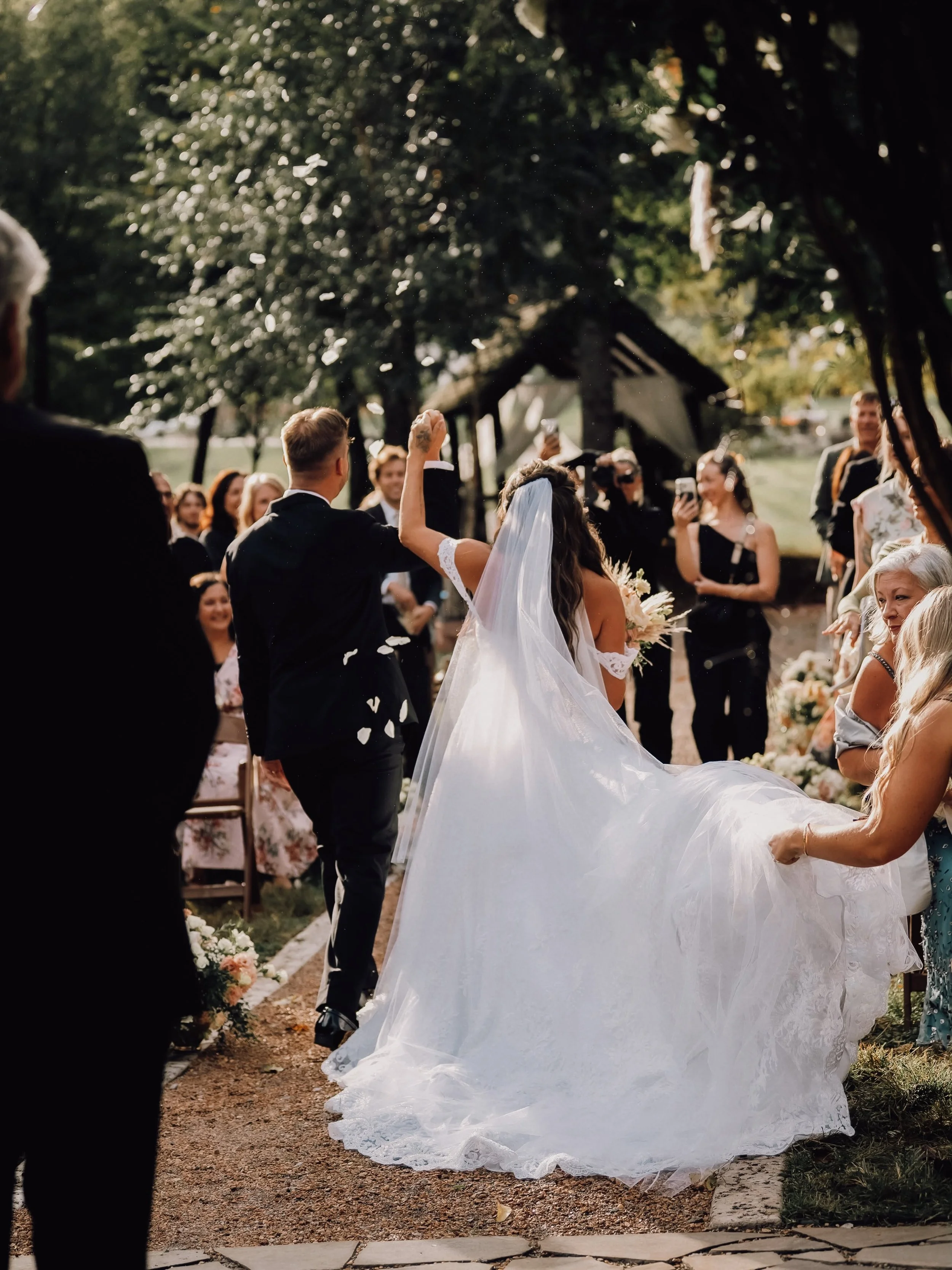 Kara Lewis and West Webb walk down the aisle with joined hands raised as guests toss petals, her lace train trailing behind at Ramble Creek.