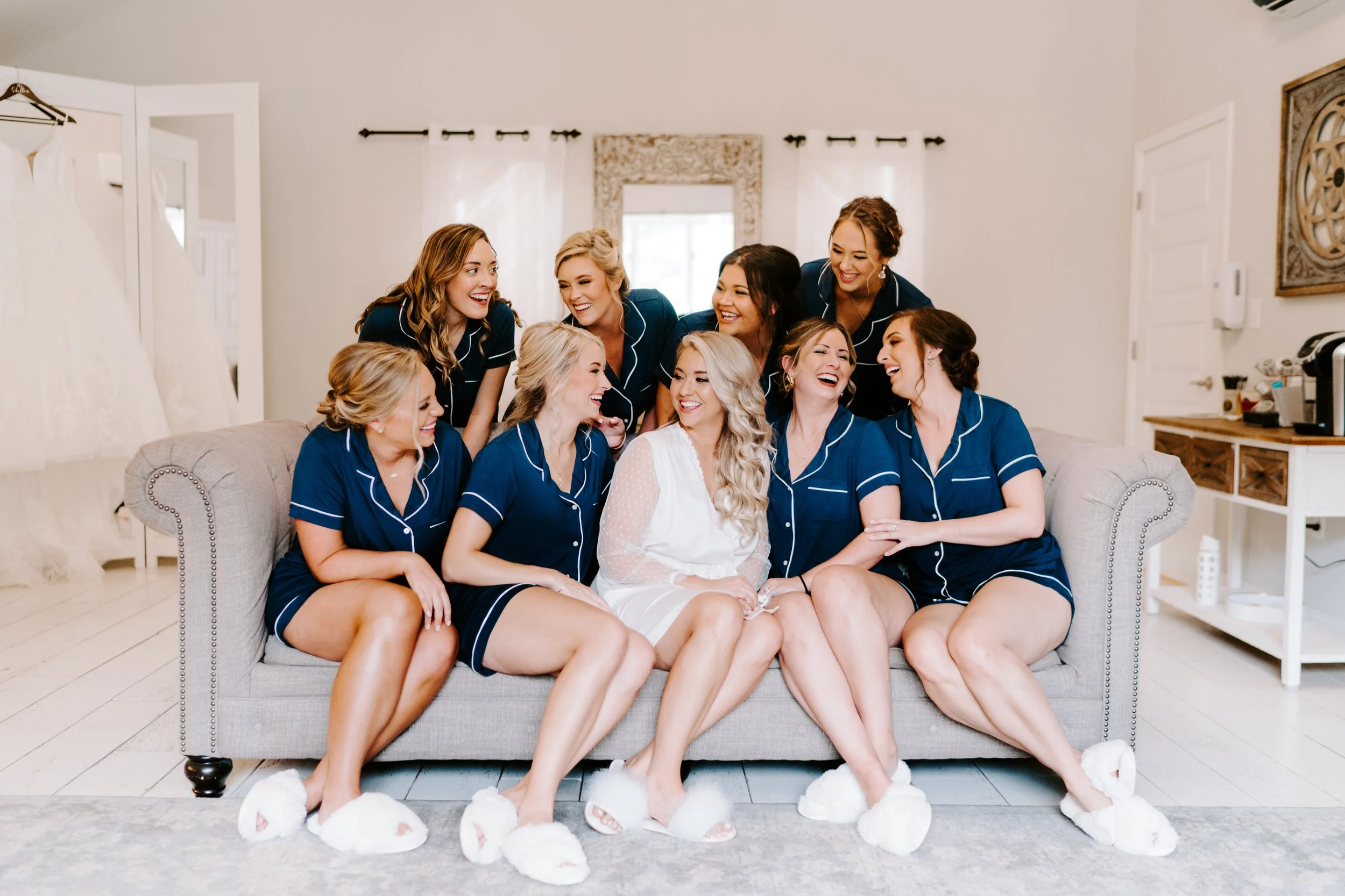 Group of bridesmaids in matching navy pajamas, some wearing white fluffy slippers, gathered in bridal suite, smiling and laughing with a bride in a white robe, celebrating a wedding or bridal occasion.