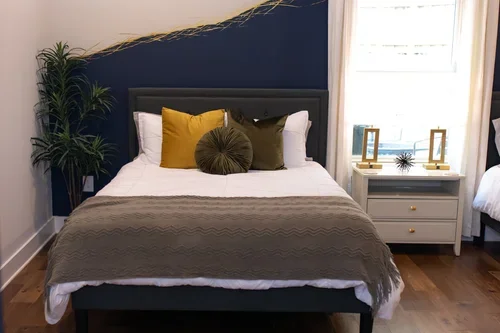 A neatly made bed with white sheets, a brown throw blanket, and multiple decorative pillows in yellow, brown, and green in a bedroom. A window with curtains and a nightstand with picture frames and a small decorative item are visible.