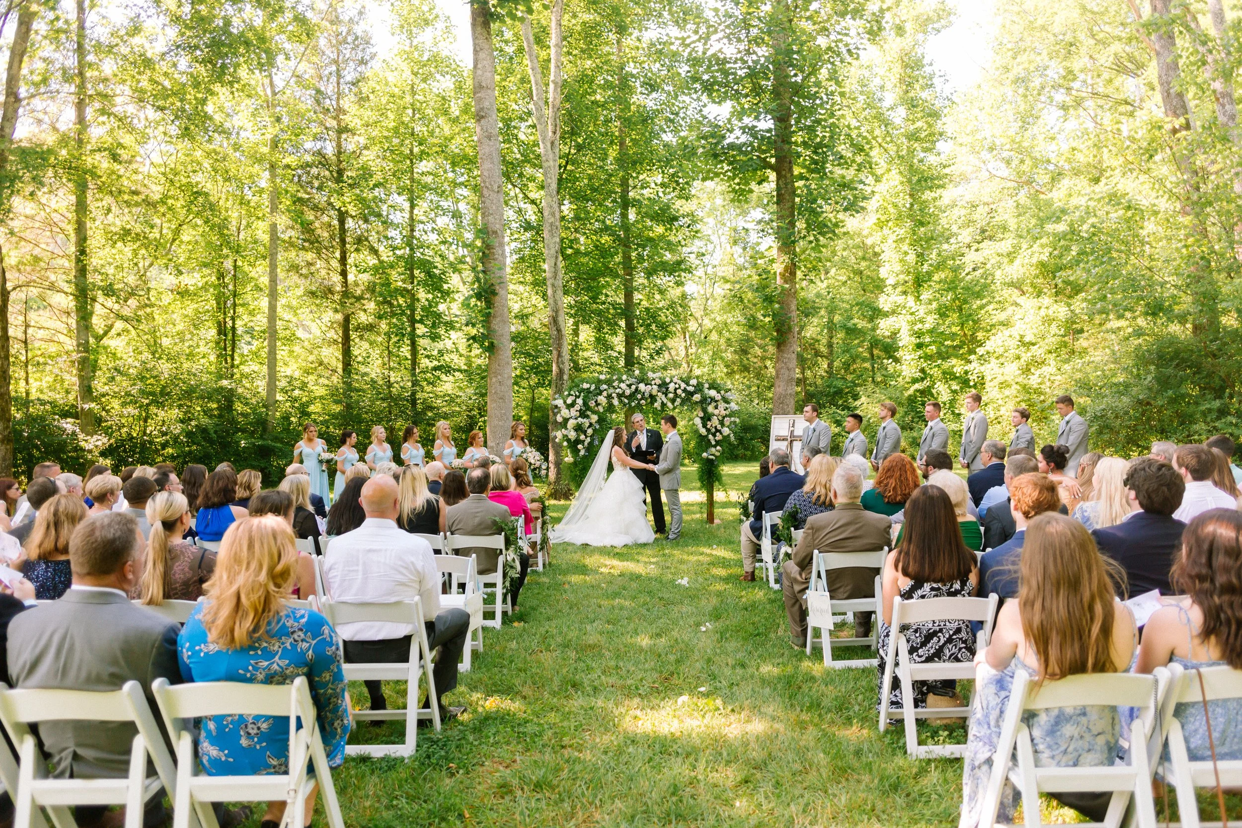 Wedding ceremony outdoors under tall trees, with bride and groom holding hands, surrounded by wedding party and guests at Ramble Creek private wedding venue.