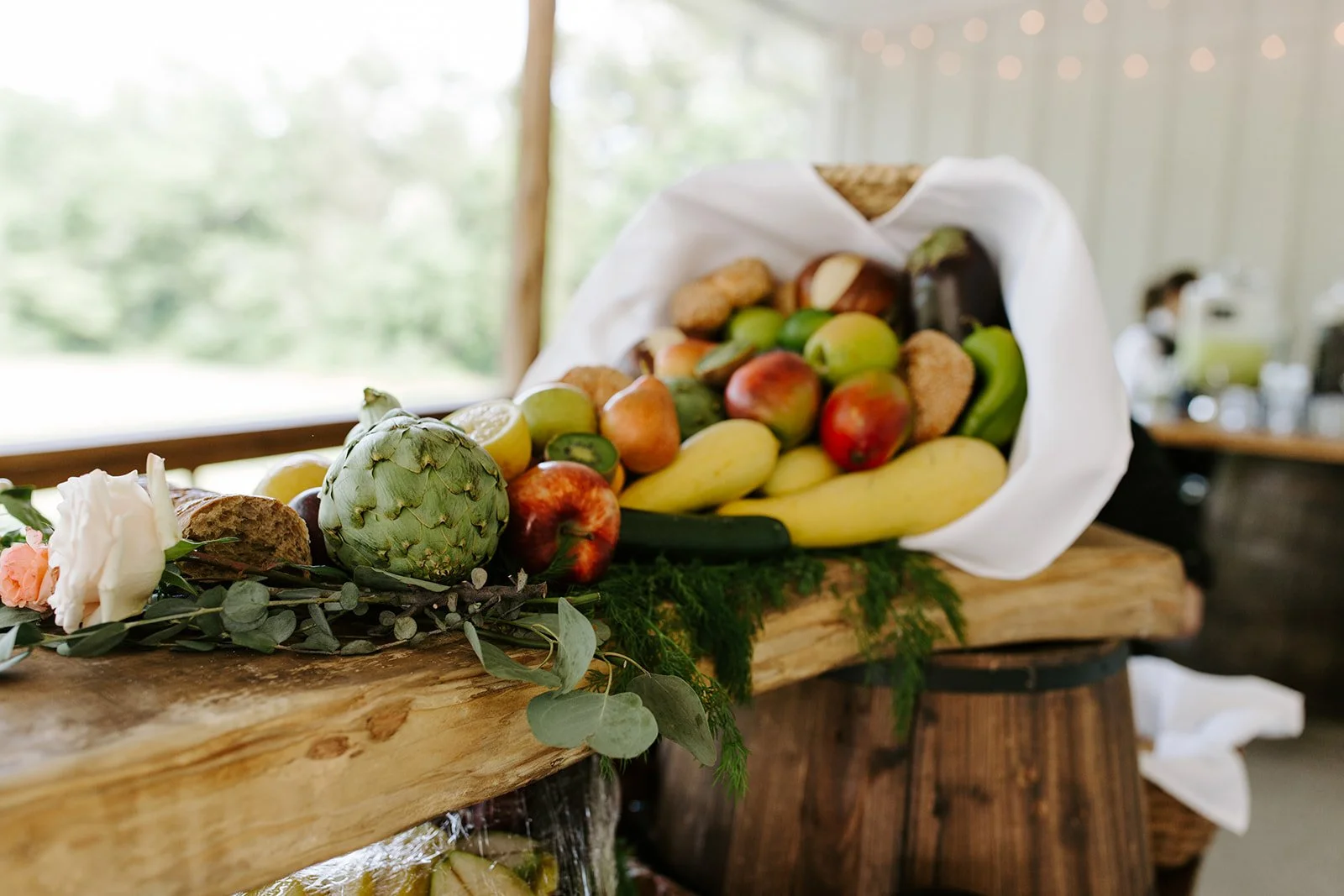 A wooden table with a white cloth lining holding a pile of assorted fresh fruits, including bananas, apples, grapes, and an artichoke, with greenery and flowers nearby. In the background, a blurred indoor cocktail hour at wedding reception.