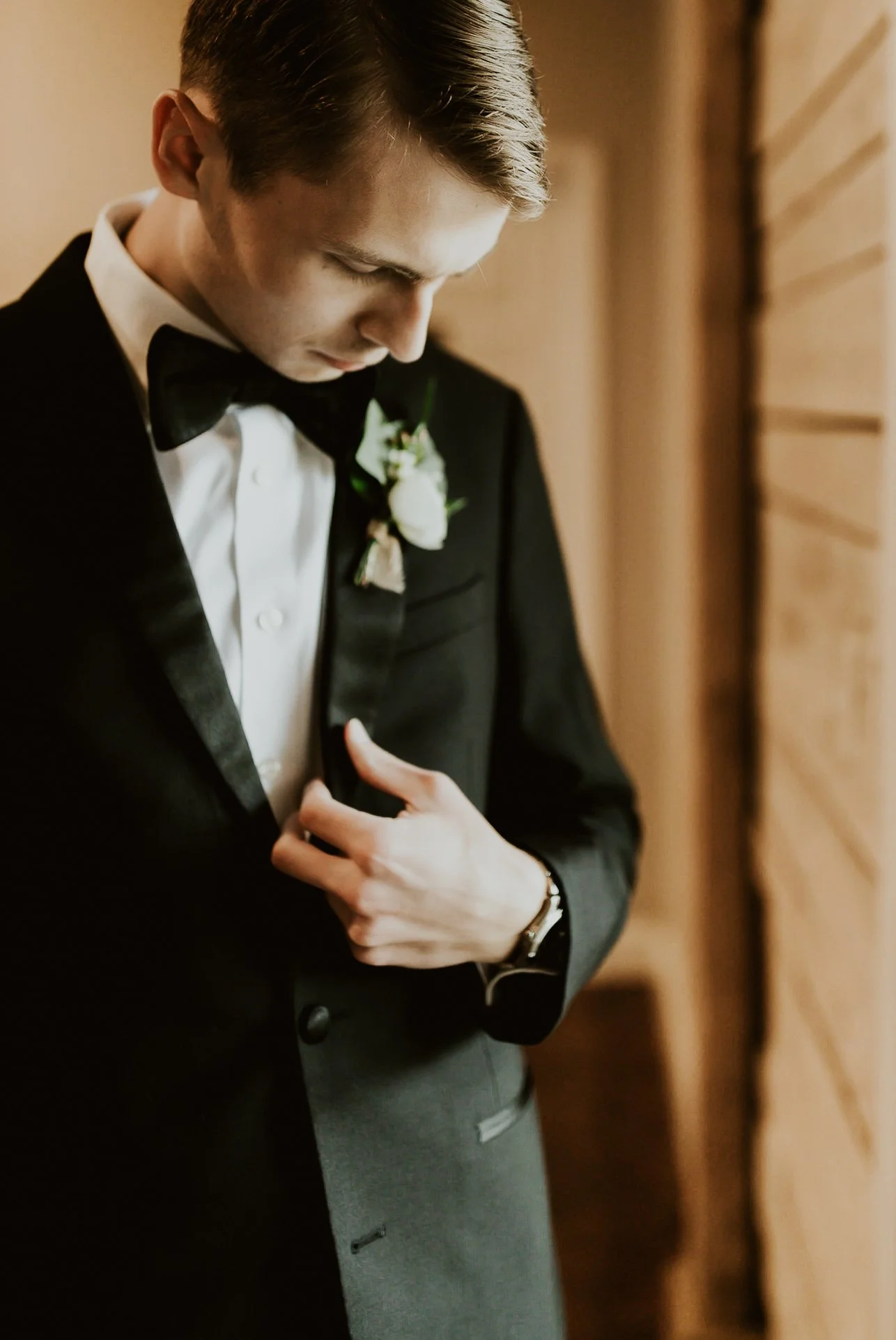 Young man in a tuxedo with a white shirt and black bow tie, standing beside a wooden wall, looking down with his hand on his chest.
