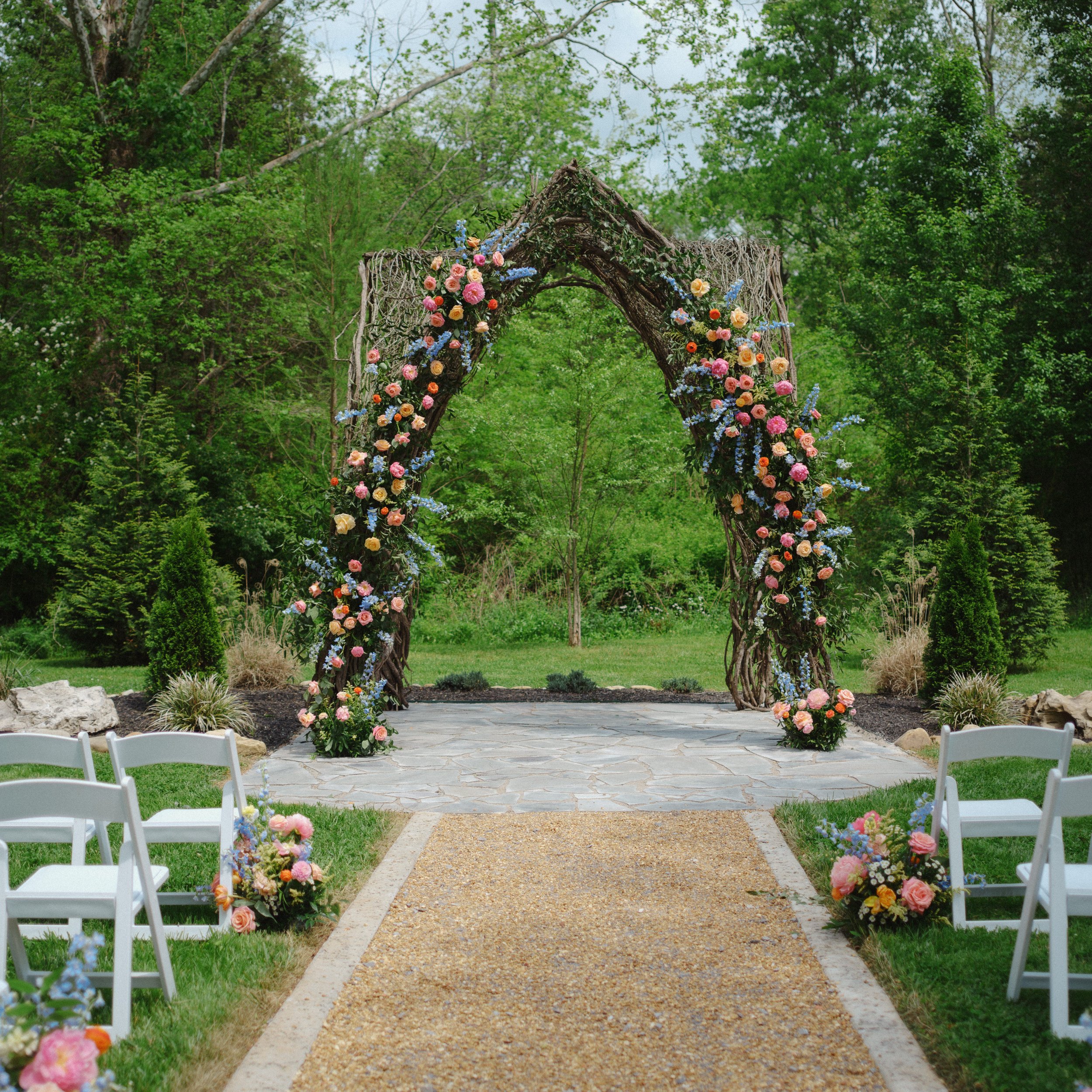 Wedding arch decorated with orange, pink, yellow, and blue flowers, set on a gravel path surrounded by white chairs and lush green trees in the background.