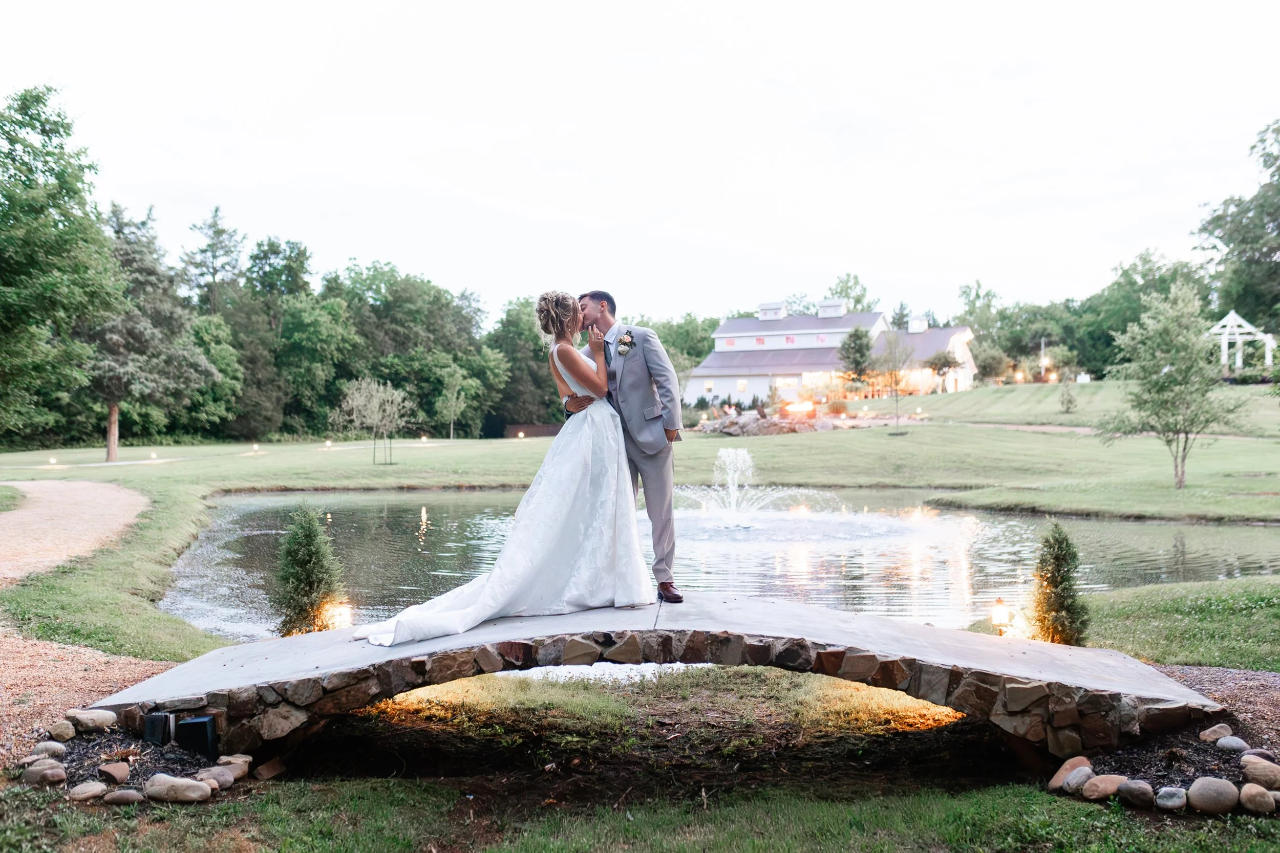Bride and groom kiss on the stone bridge by the pond at Ramble Creek with the venue glowing in the background.