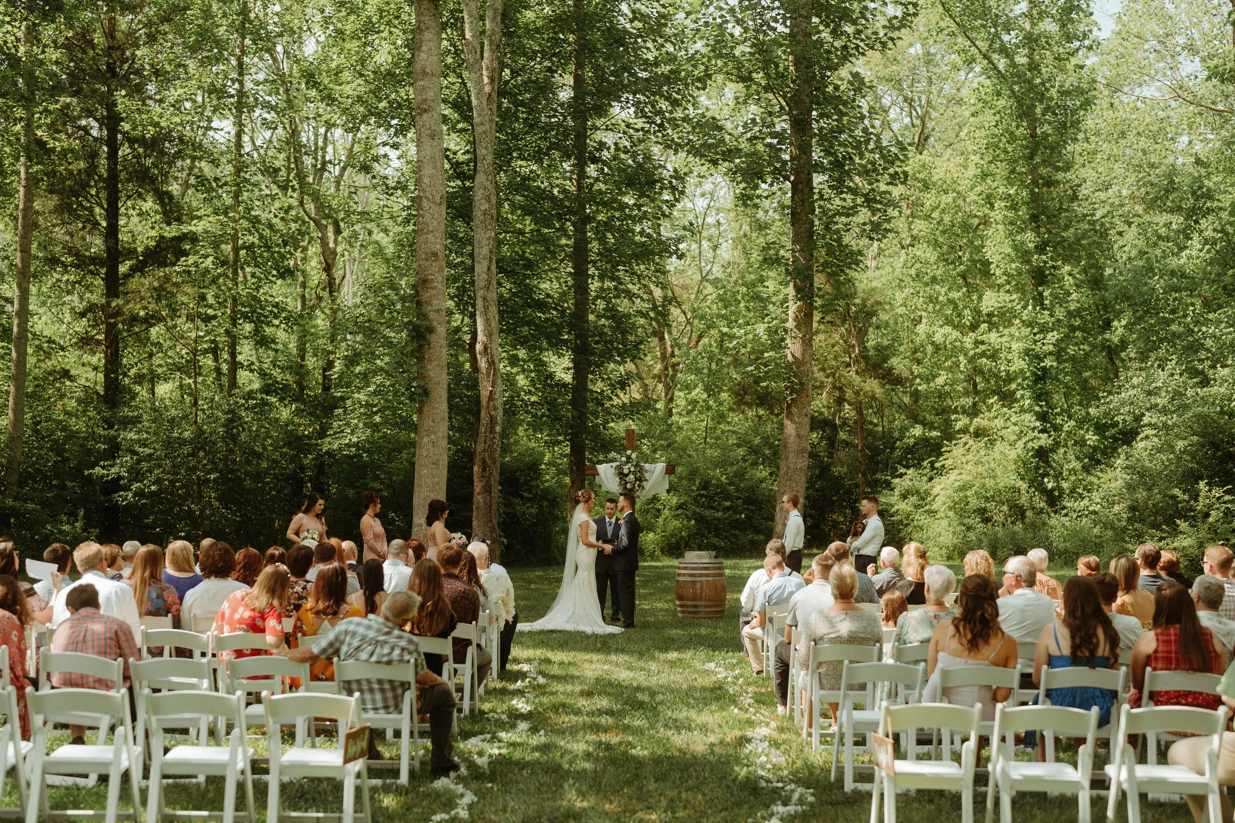 Outdoor wedding ceremony in a wooded area at Ramble Creek all-inclusive wedding venue with guests seated on white chairs, a couple exchanging vows at the altar, surrounded by tall trees and sunlight filtering through the leaves.