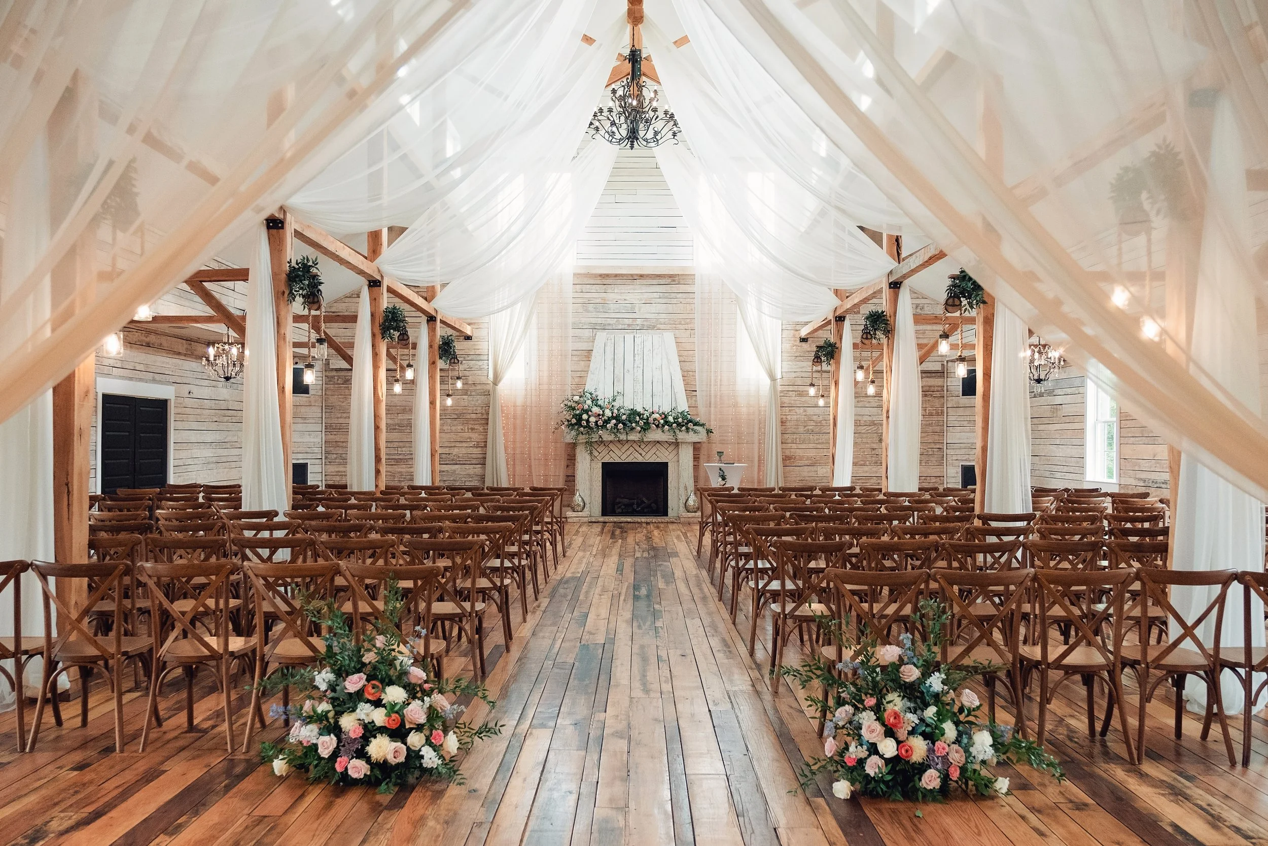 Indoor wedding ceremony setup in a bright wooden grand hall, with rows of cross-back chairs, white draped fabric overhead, floral arrangements lining the aisle, and a fireplace altar decorated with greenery and pastel flowers