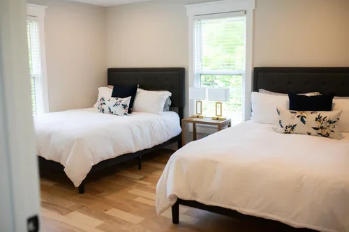 Bedroom inside the Villa at Ramble Creek, featuring two queen beds with white bedding, dark headboards, floral accent pillows, a shared nightstand, and bright windows.