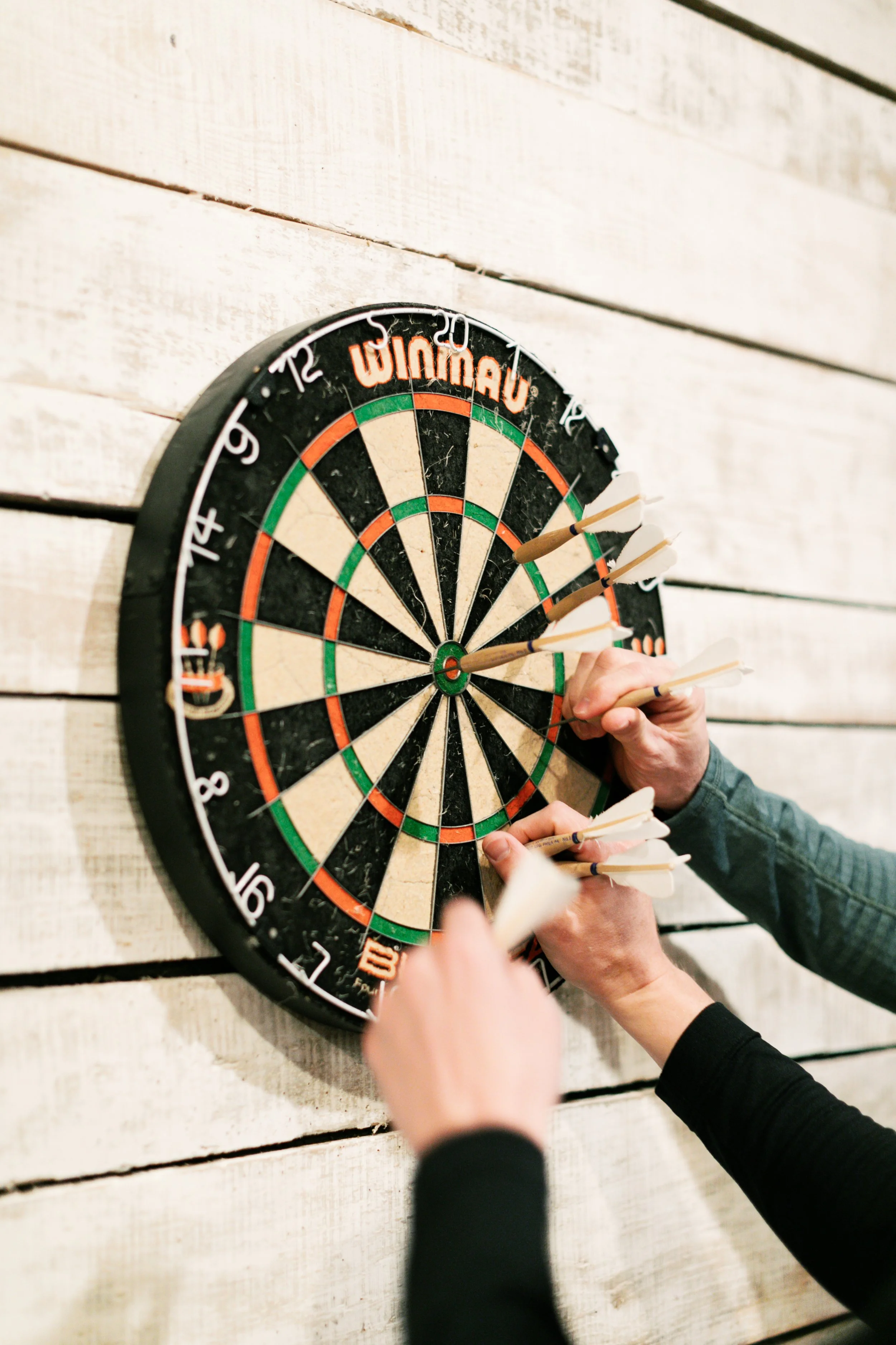 Multiple people throw darts at a dartboard attached to a white wooden wall in the groom's quarters at Ramble Creek, a private East TN destination wedding venue.