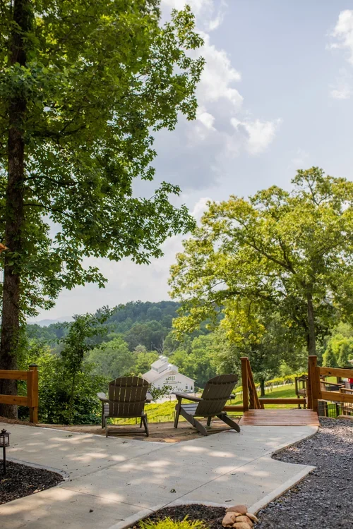 Two Adirondack chairs on a wooden deck overlooking a lush green landscape with trees and distant hills under a partly cloudy sky. One of three accommodations at Ramble Creek Tennessee wedding venue with on-site lodging.