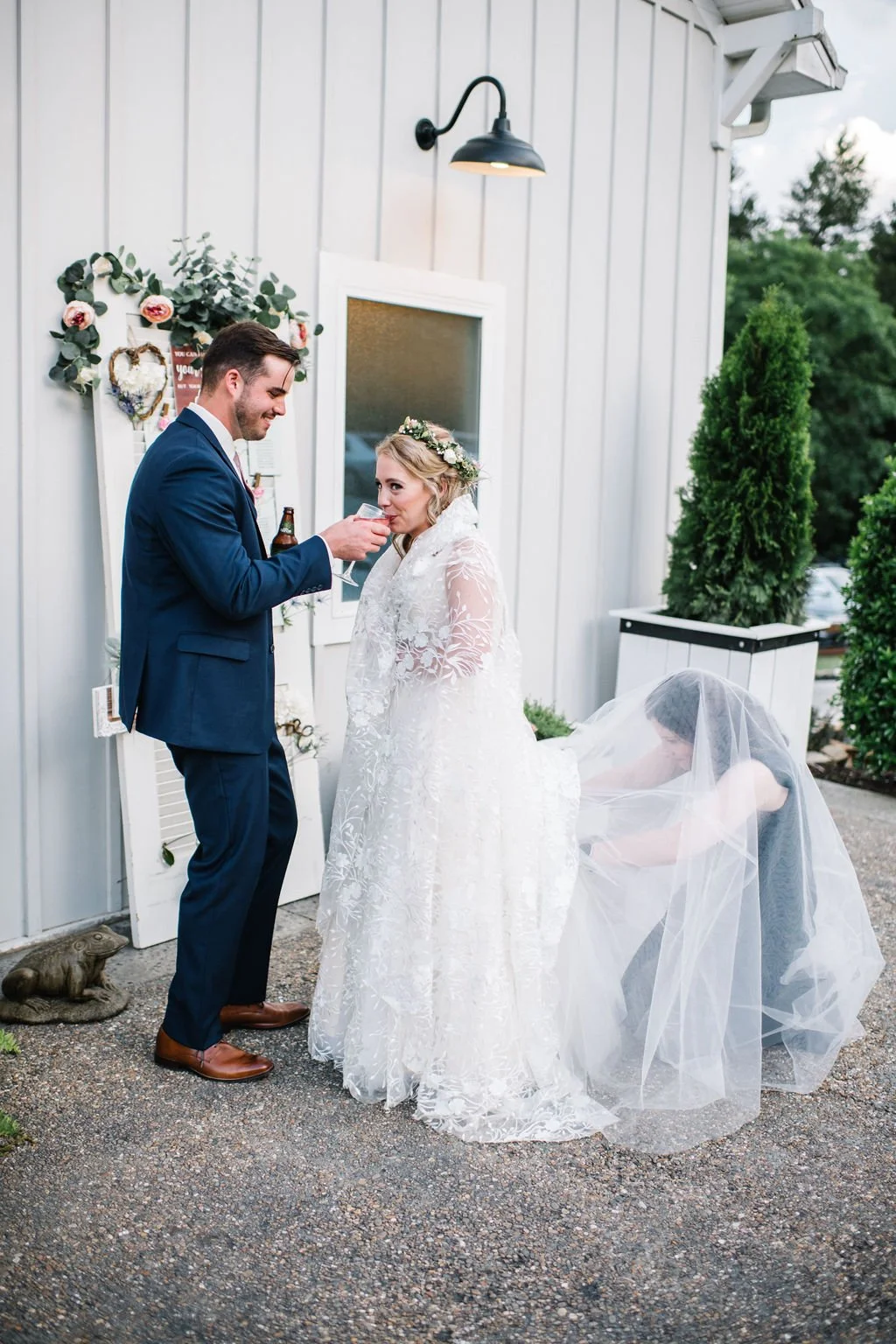 Ramble Creek wedding coordinator helping arrange the bride’s veil as the bride and groom share a drink outside the white wedding venue exterior