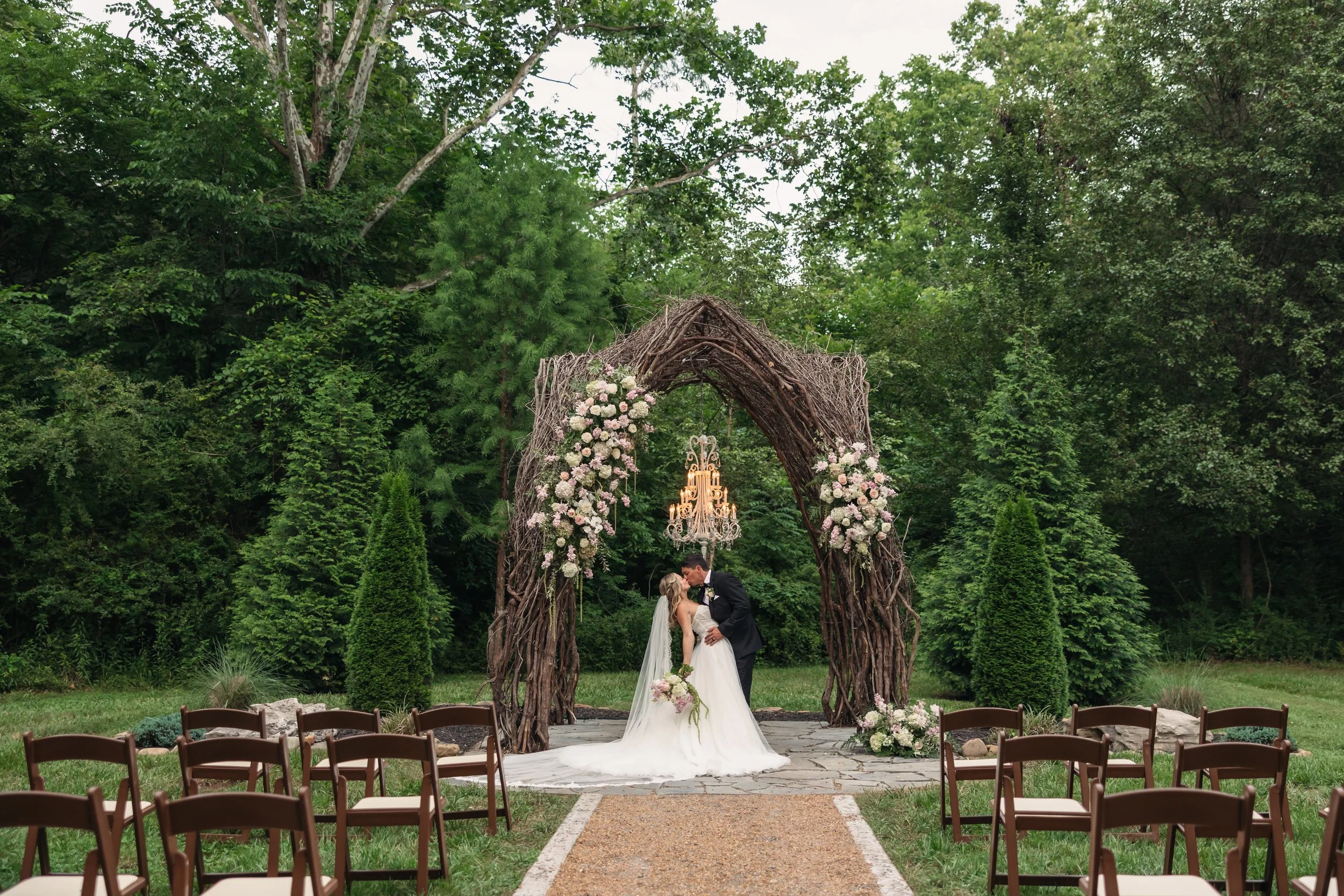 Bride and groom kissing beneath the outdoor ceremony arch at Ramble Creek with floral arrangements, chandelier lighting, wooden chairs, and a woodland backdrop.