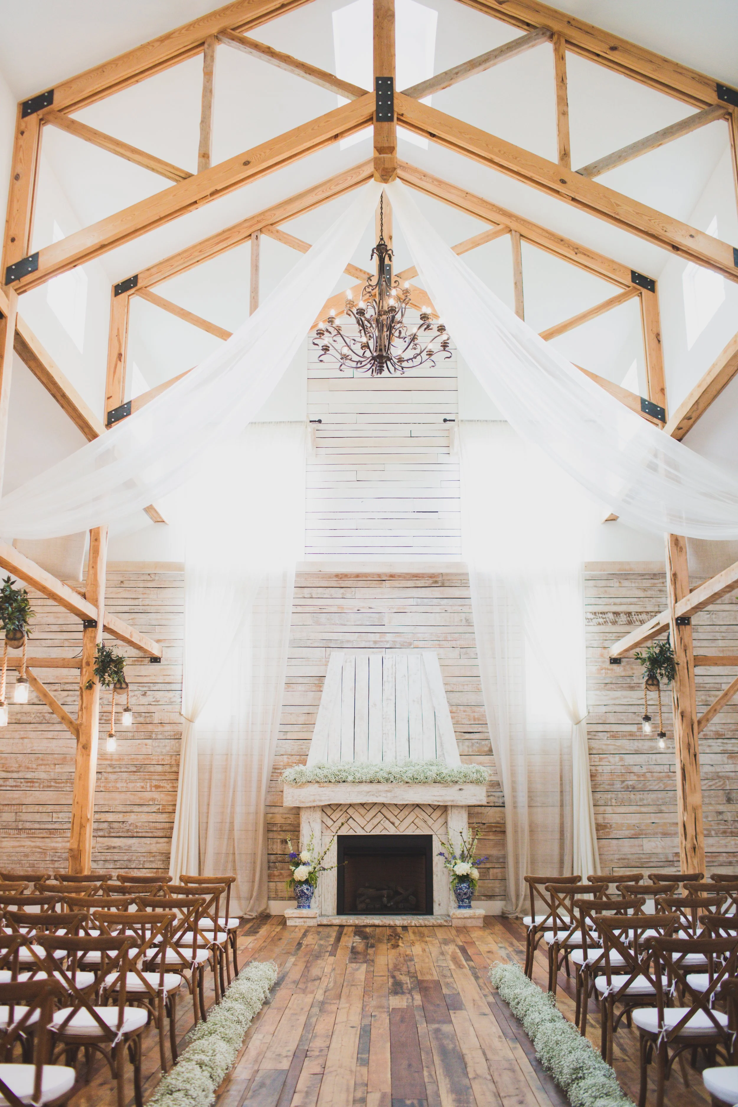 Best indoor wedding ceremony space in Tennessee with wooden beams, a chandelier, white drapes, a fireplace, floral arrangements, and rows of chairs facing the altar.