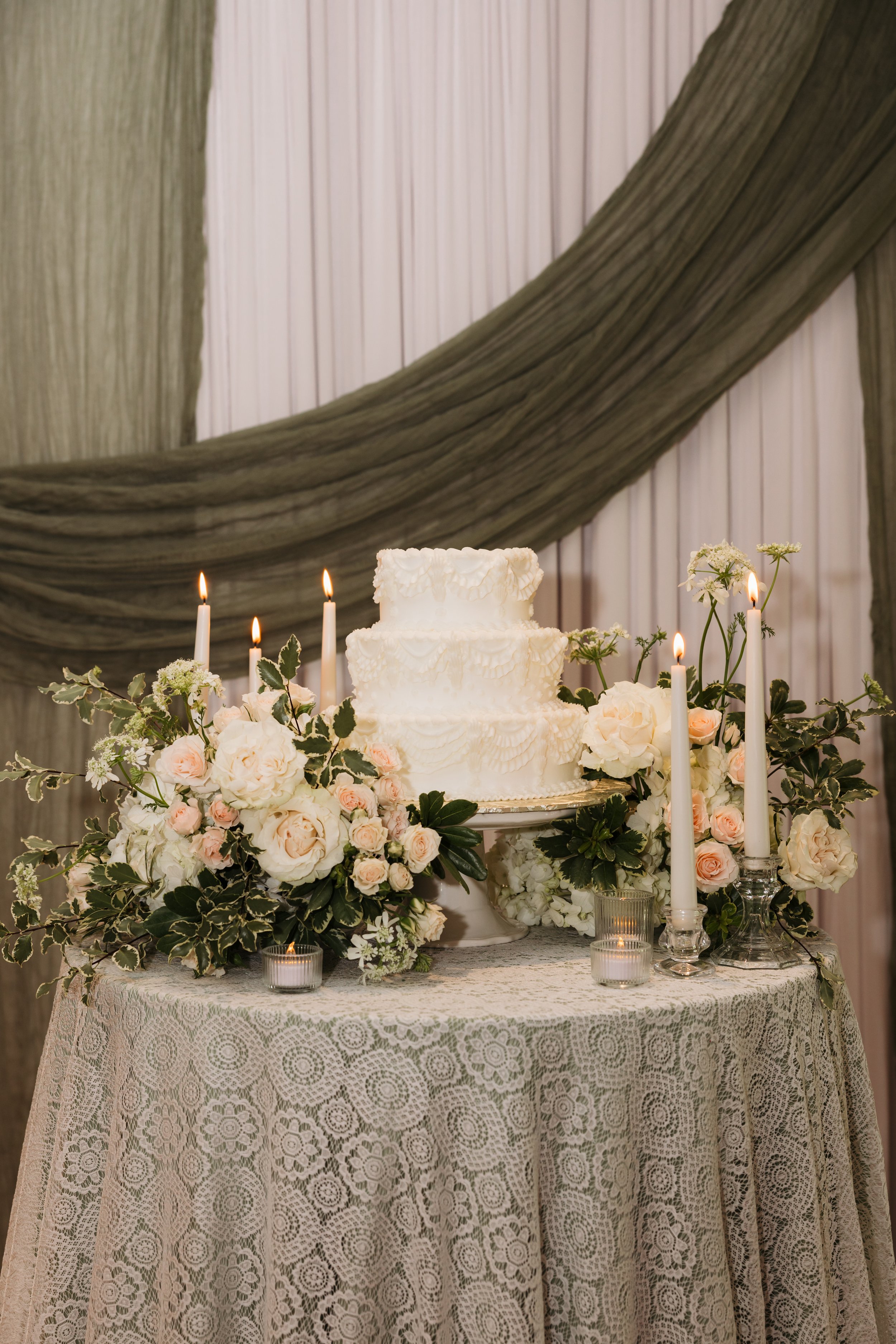 White wedding cake displayed at Ramble Creek with soft floral arrangements, candlelight, lace linens, and elegant green draping
