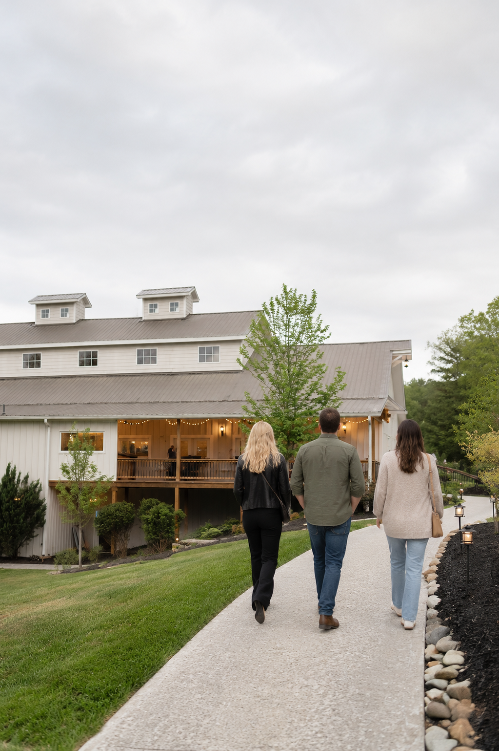 Three people walk along the pathway toward Ramble Creek during a daytime property tour, with the venue and landscaped grounds softly glowing in the background