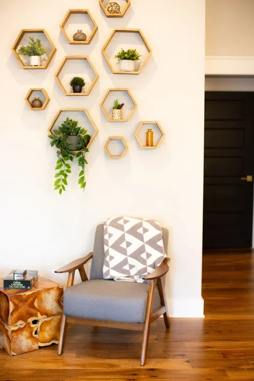 Wall-mounted hexagonal wooden shelves with potted plants and decorative objects above a gray armchair with a patterned throw blanket, next to a small wooden side table, in a room with hardwood floors and a black door.