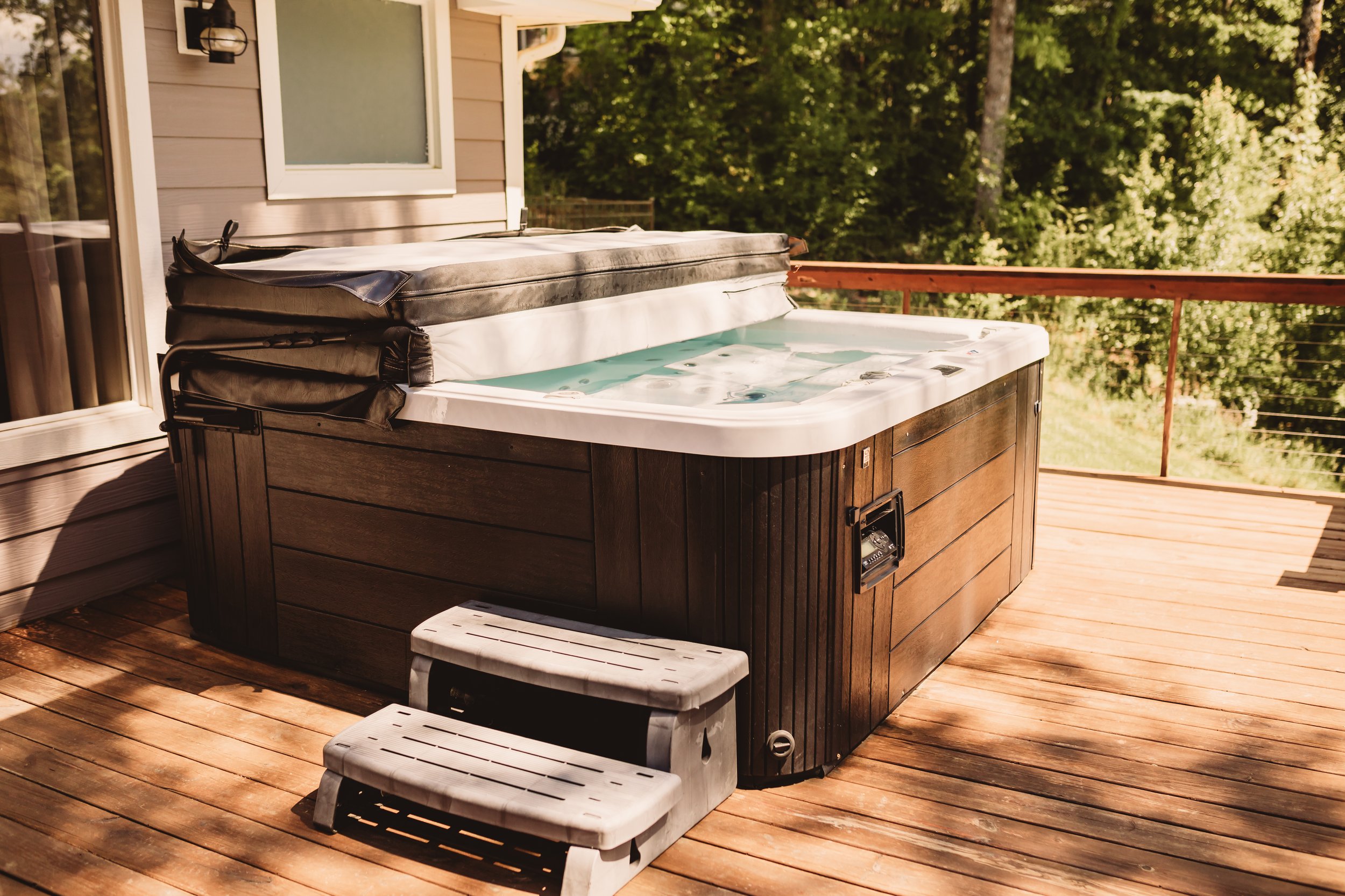 Wooden deck with hot tub and small white stairs outside a house with beige siding and a frosted window. Green trees in the background.