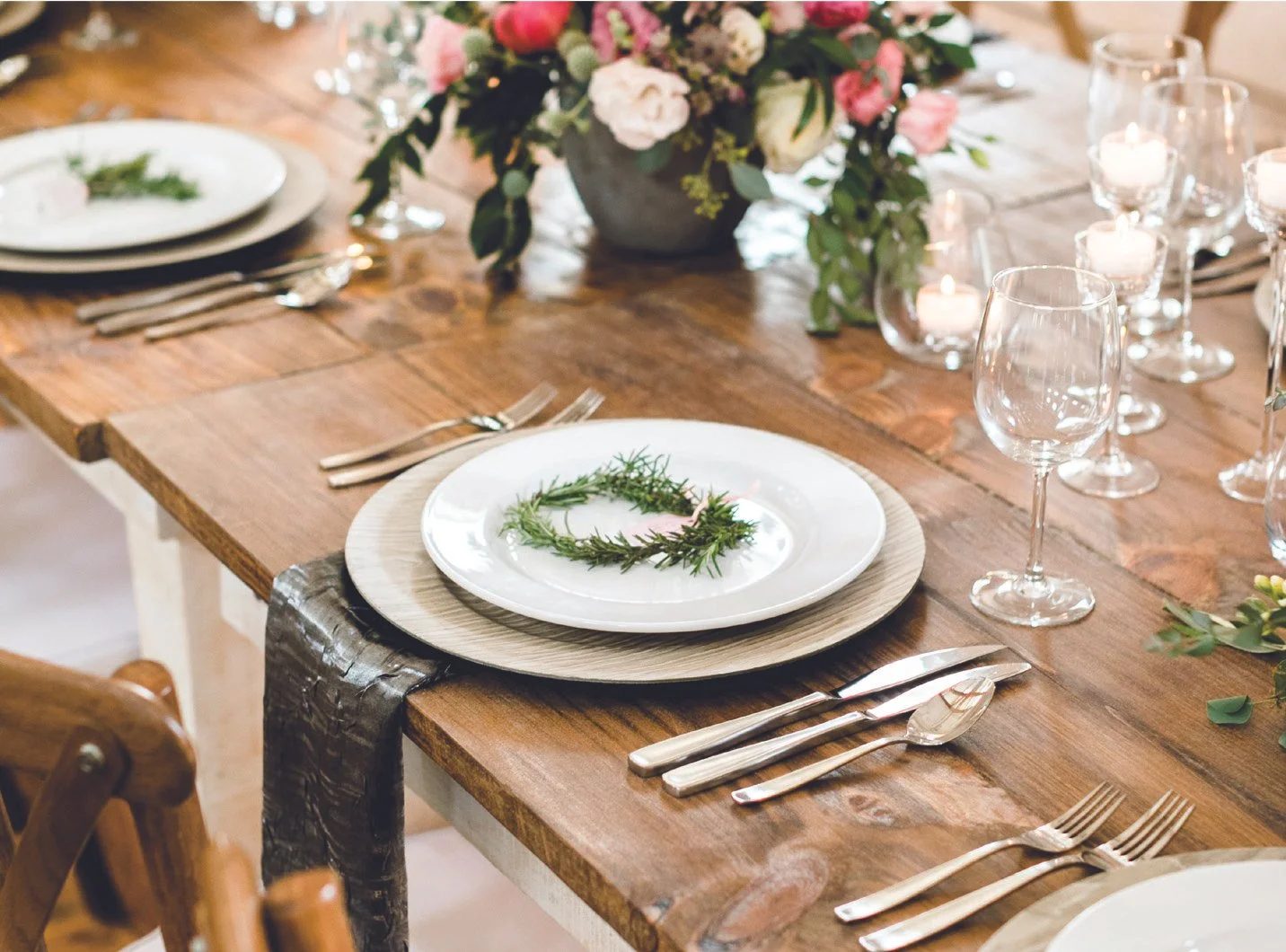 A rustic wooden dining table set for a wedding with white plates, silverware, wine glasses, and a centerpiece of candles and a floral arrangement. One place setting has a green wreath on the plate.