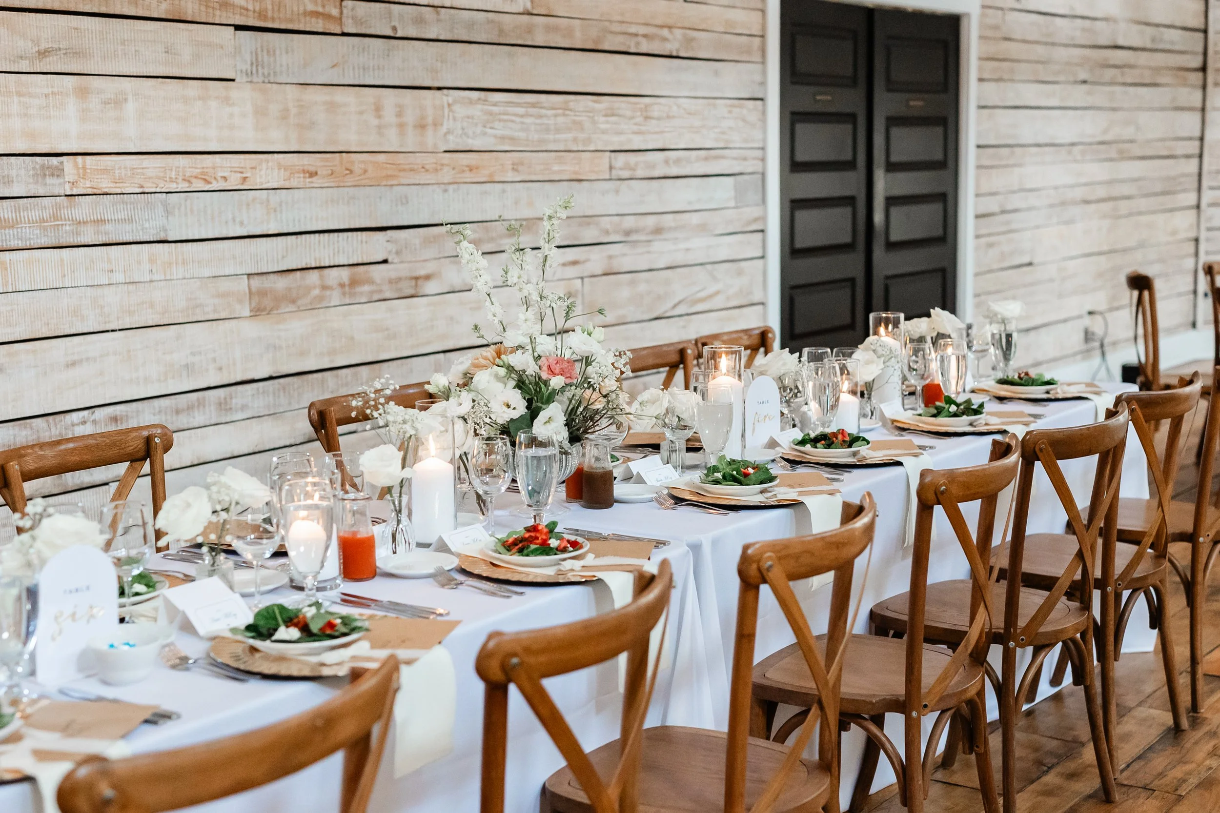 Ramble Creek wedding reception tables with white linens, wood cross-back chairs, candlelit floral centerpieces, gold place settings, and elegant indoor décor.