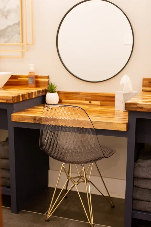 A modern bathroom vanity with a wooden countertop, a round mirror, a transparent chair with gold legs, a small potted plant, a soap dispenser, and a box of tissues. One of three homes at Ramble Creek Tennessee wedding venue with on-site accommodation