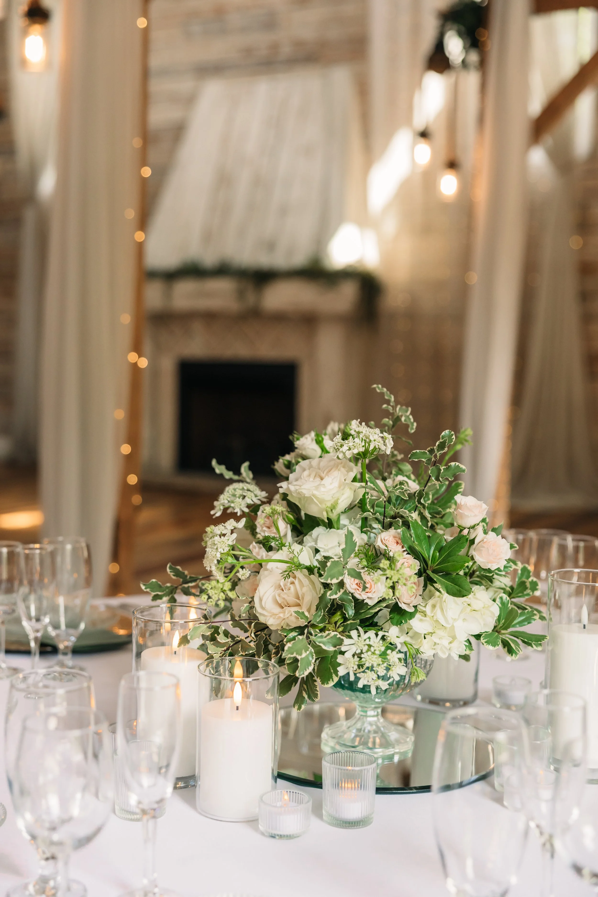 Romantic wedding centerpiece floral arrangement with white roses, soft blush blooms, greenery, and candlelight on a reception table at Ramble Creek wedding venue in Tennessee.