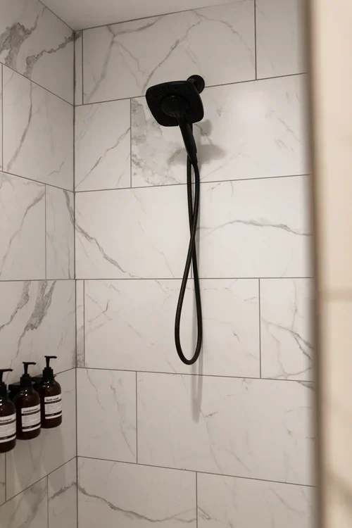 Bathroom shower inside the Villa at Ramble Creek, featuring marble-style tile, black shower fixtures, and built-in shelving with bath products.