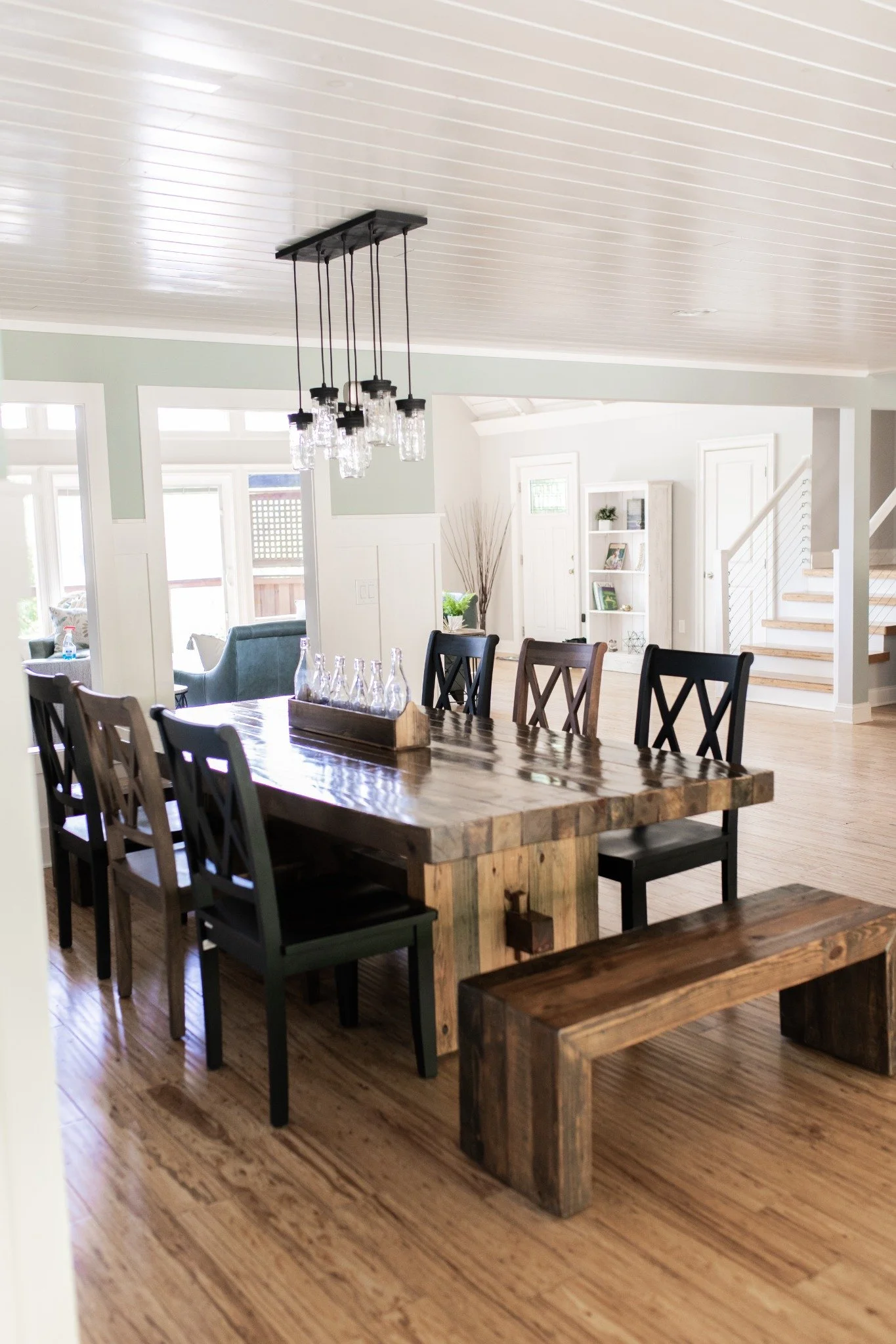 Dining room at the Manor onsite accommodation at Ramble Creek, featuring a rustic wooden table, mixed seating, modern chandelier, warm wood floors, and an open view into the living area.