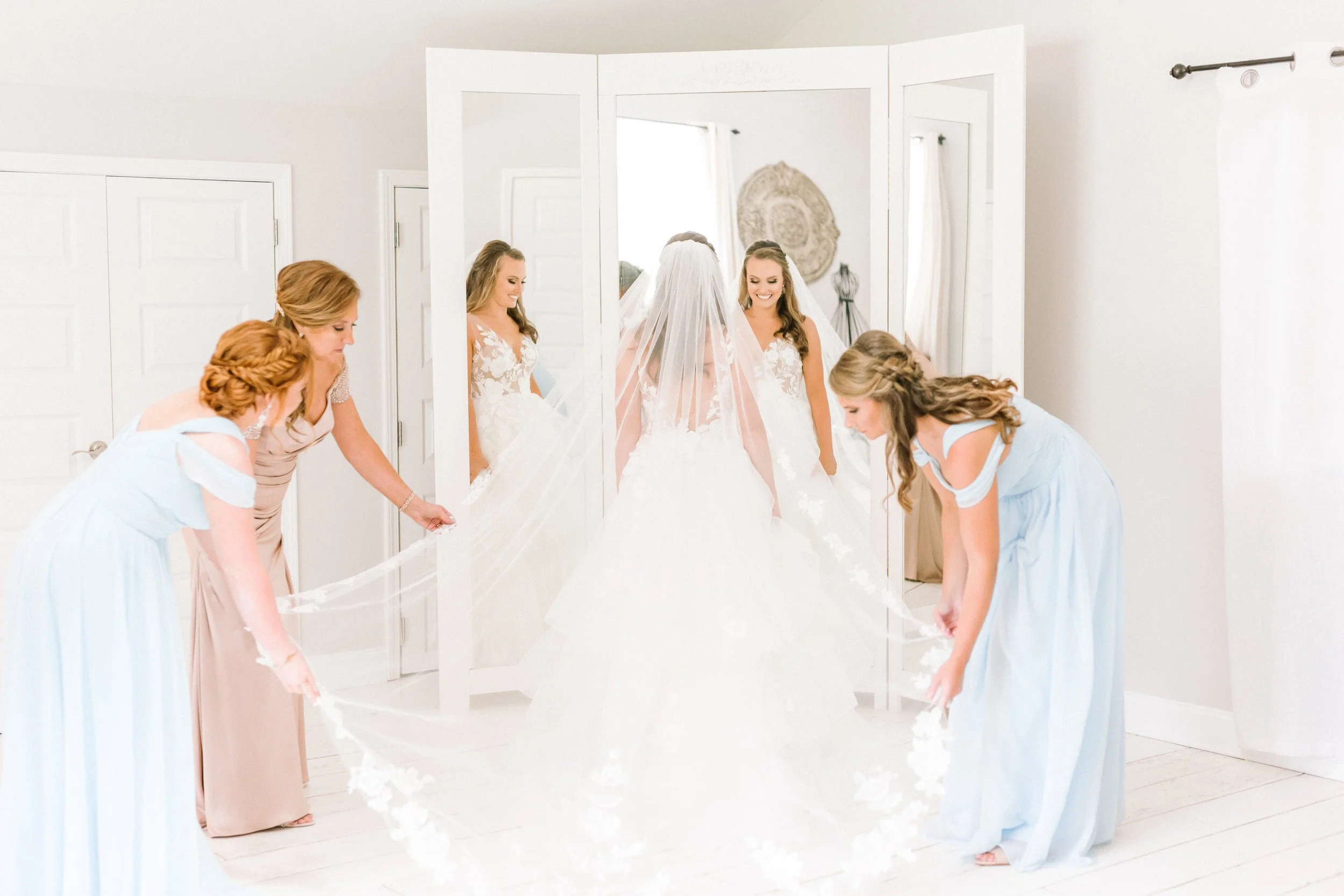 Bridesmaids helping bride with her wedding dress in front of a full-length mirror, in a bright room at Ramble Creek Tennessee wedding venue with in-house hair and makeup services.