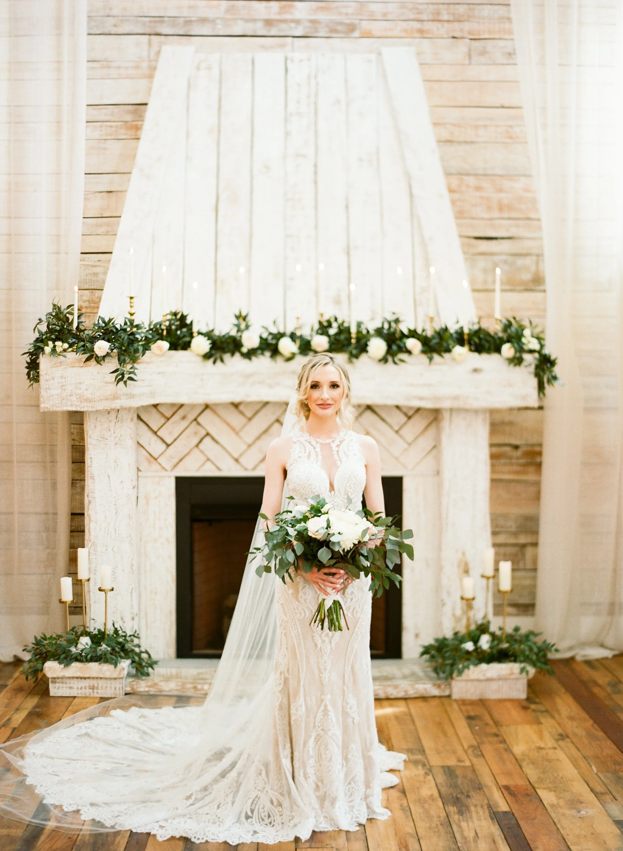 A bride in a lace wedding dress holding a bouquet of white flowers and greenery, standing in front of a rustic fireplace with floral and candle decorations at an indoor wedding ceremony in Tennessee.